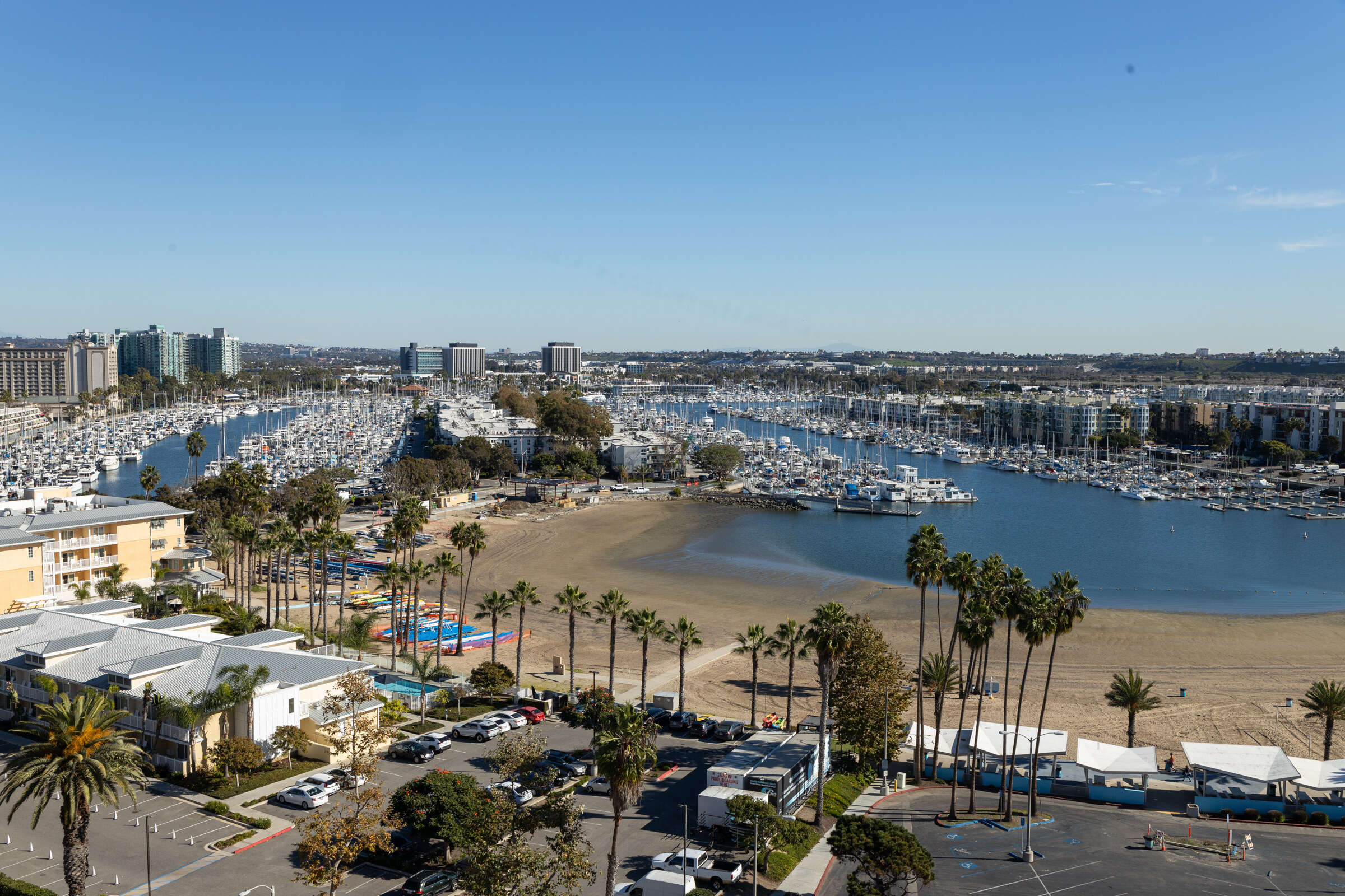 A view over a marina packed with boats, palm trees, a sandy beach and a blue sky.