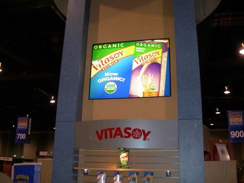 A Vitasoy sign on a tall booth structure under a screen showing organic Vitasoy soymilk cartons, with a small shelf of cartons below.