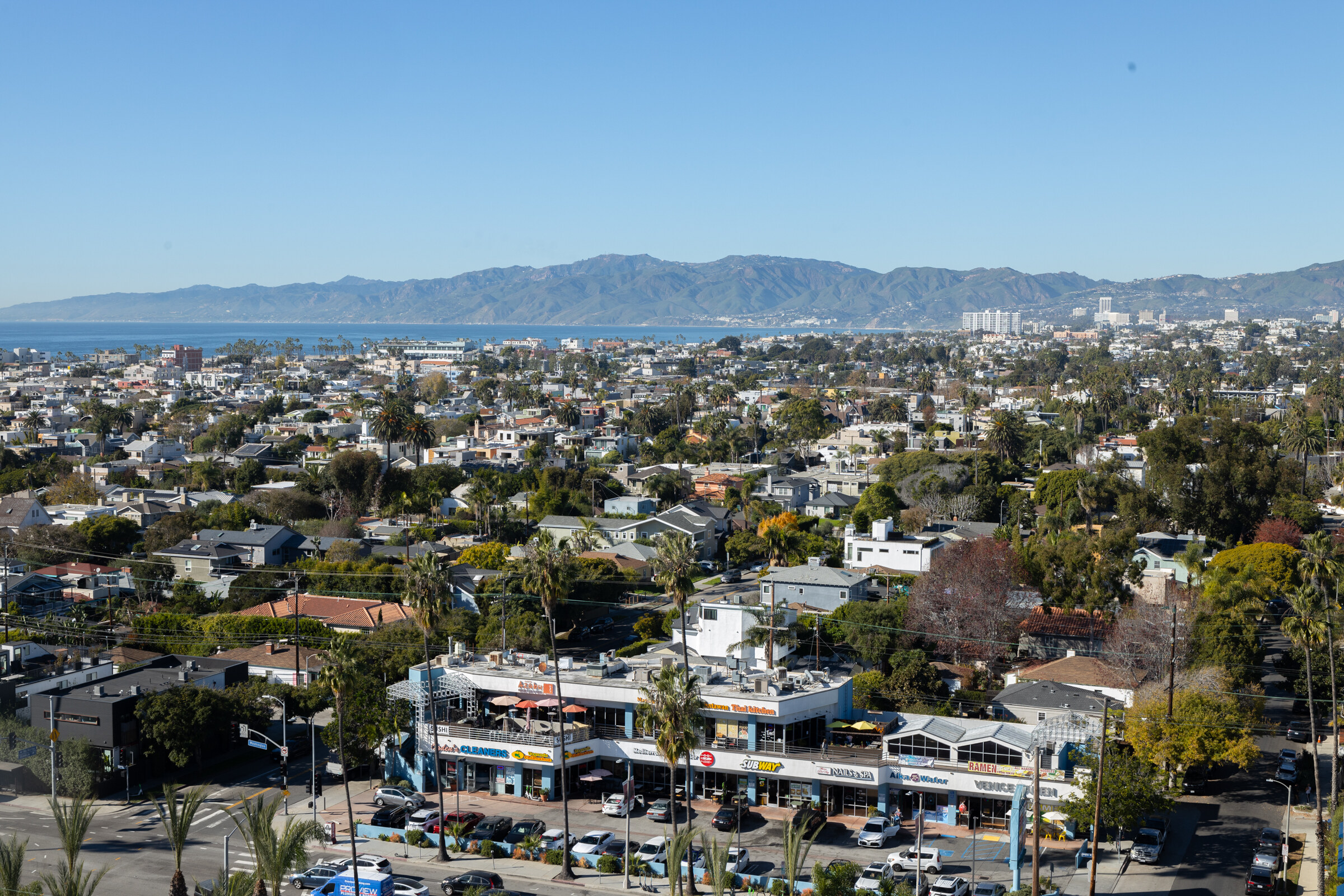 A view over rooftops and trees toward the ocean and distant mountains under a blue sky.