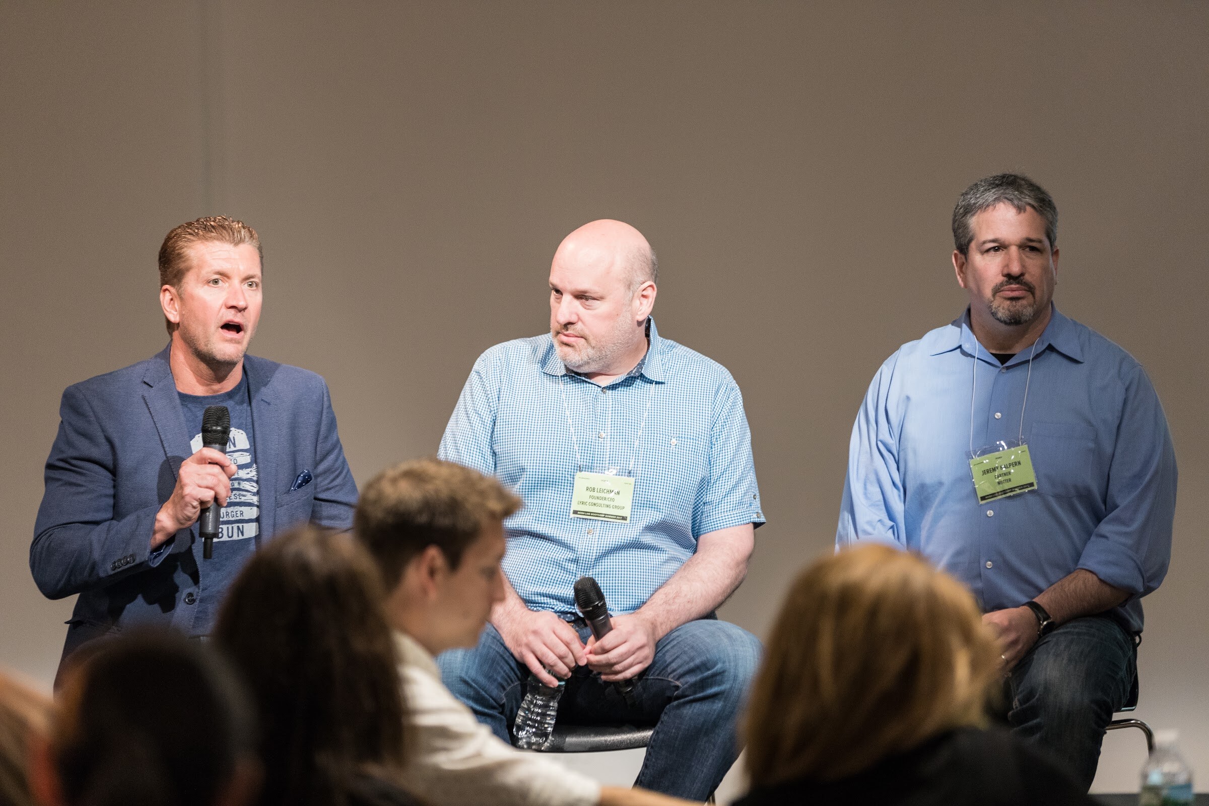 Three men on stools hold microphones and speak to a seated audience; one in a blue blazer is mid-sentence.