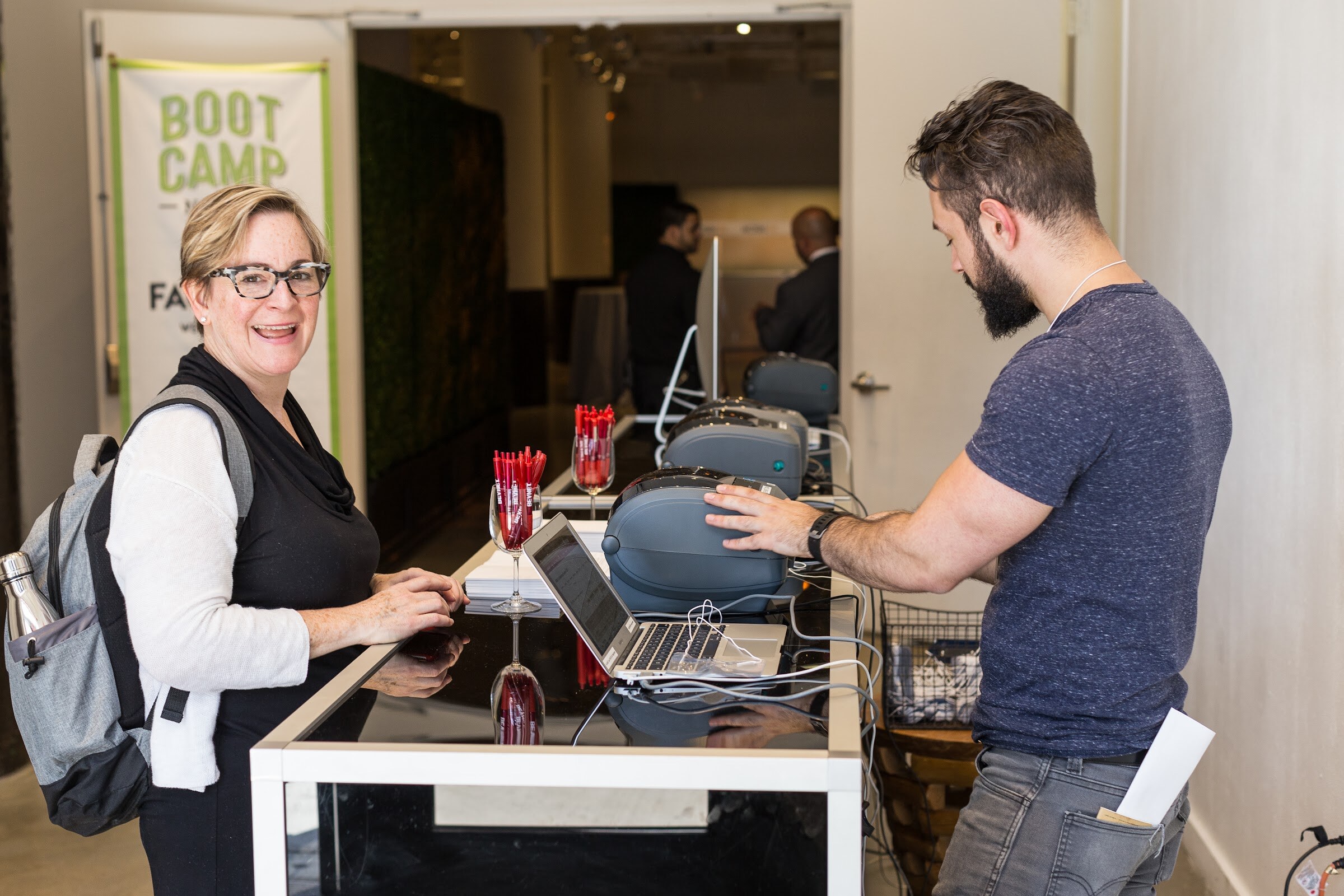 A smiling woman with a backpack stands at a registration table while a man in a navy t-shirt operates a laptop and label printers, with a Boot Camp banner behind them.