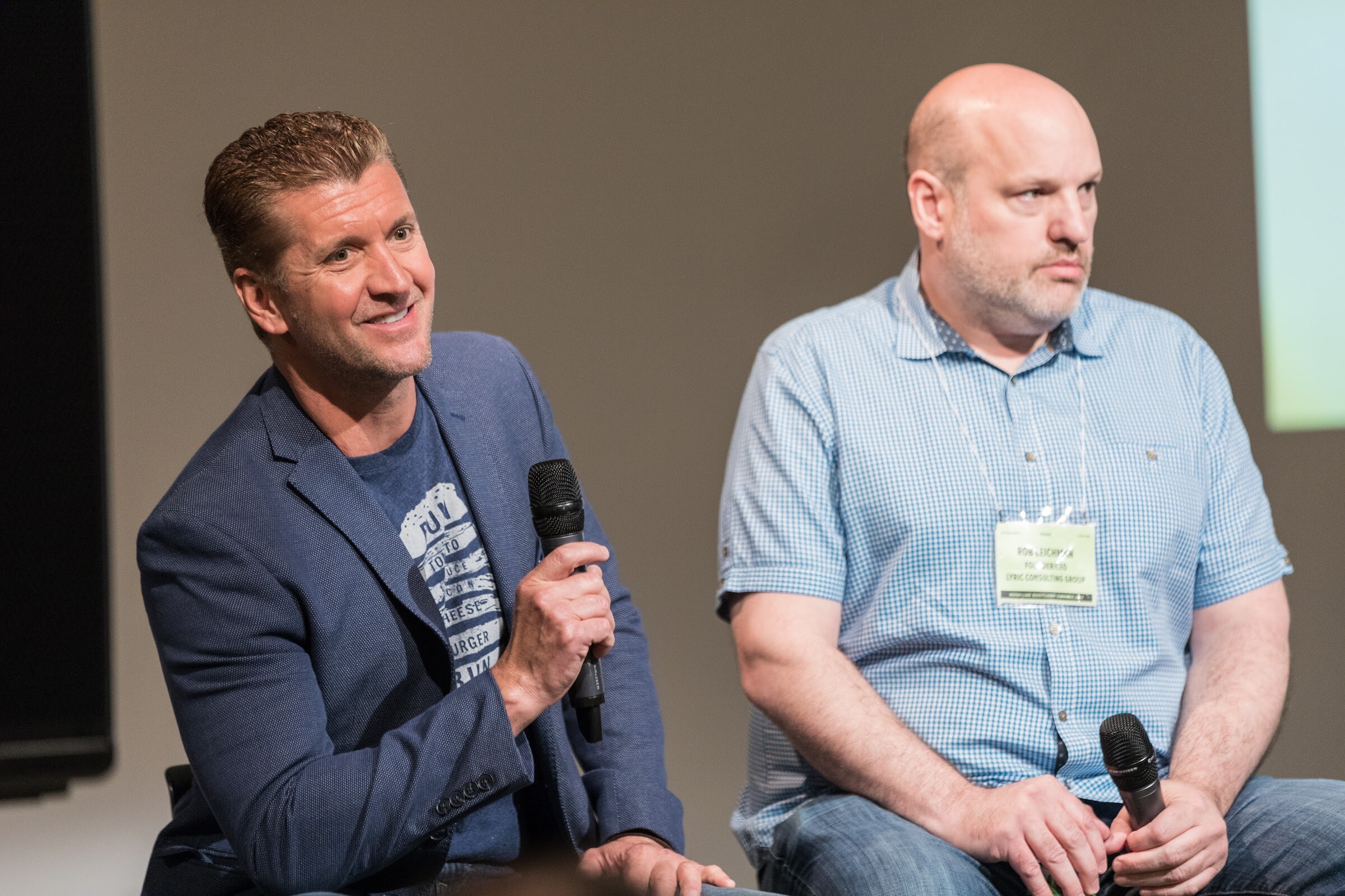A man in a blue blazer smiles and holds a microphone beside a seated man in a light blue checked shirt with a conference badge.