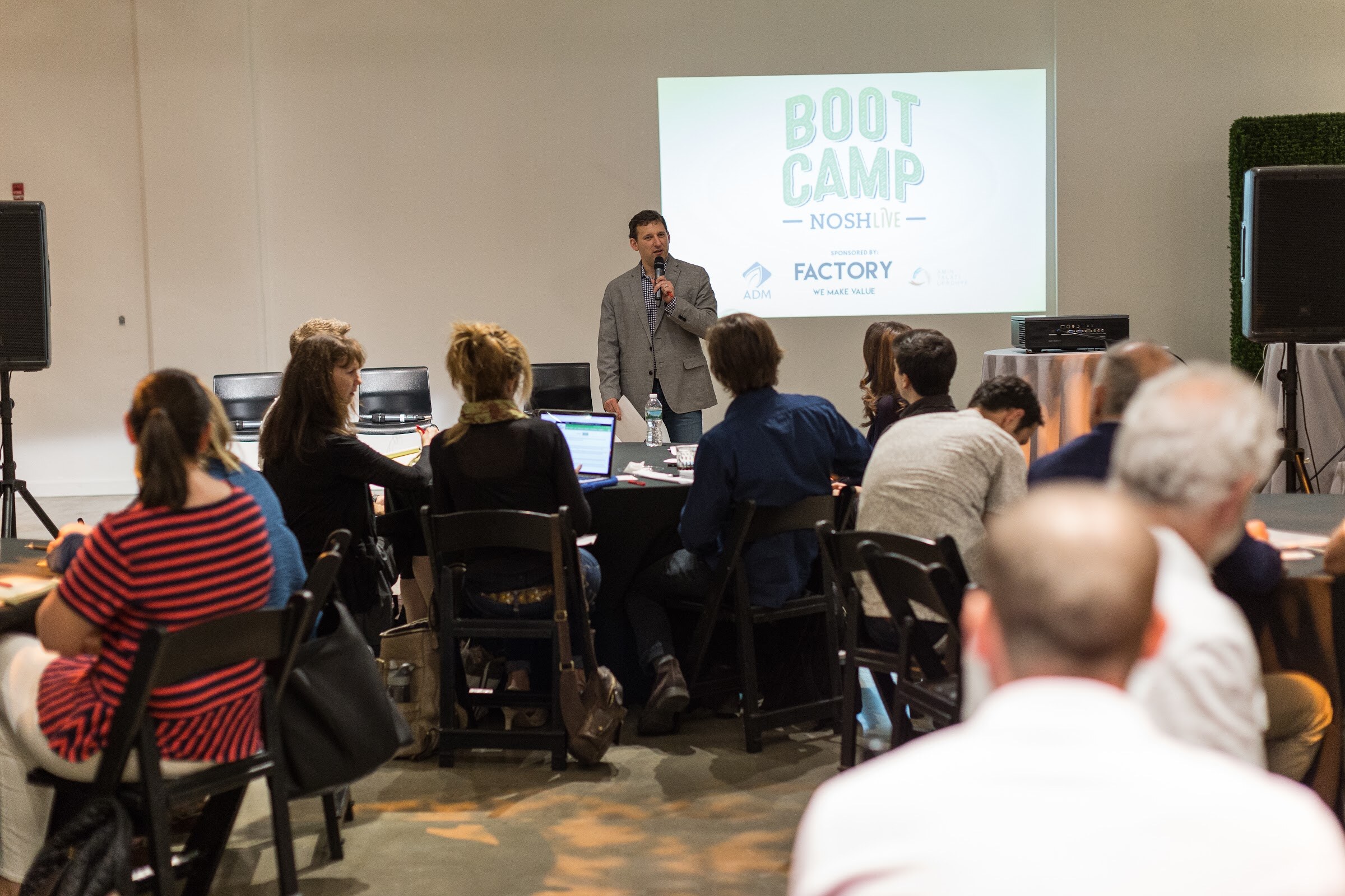 A man in a gray blazer speaks into a microphone at the front of a room of round tables, with a projected Boot Camp Nosh Live slide sponsored by Factory behind him.