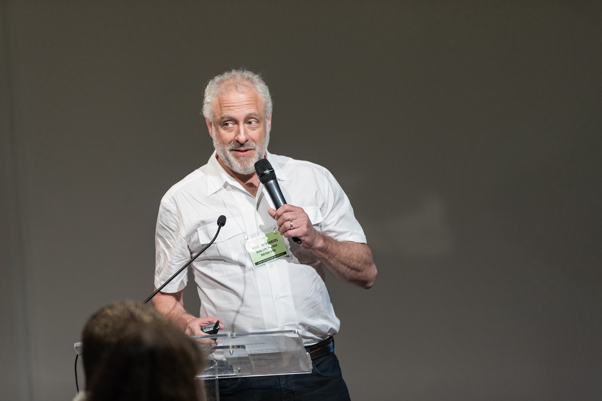 A gray-haired bearded man in a white shirt holds a handheld microphone beside a clear lectern against a gray wall.