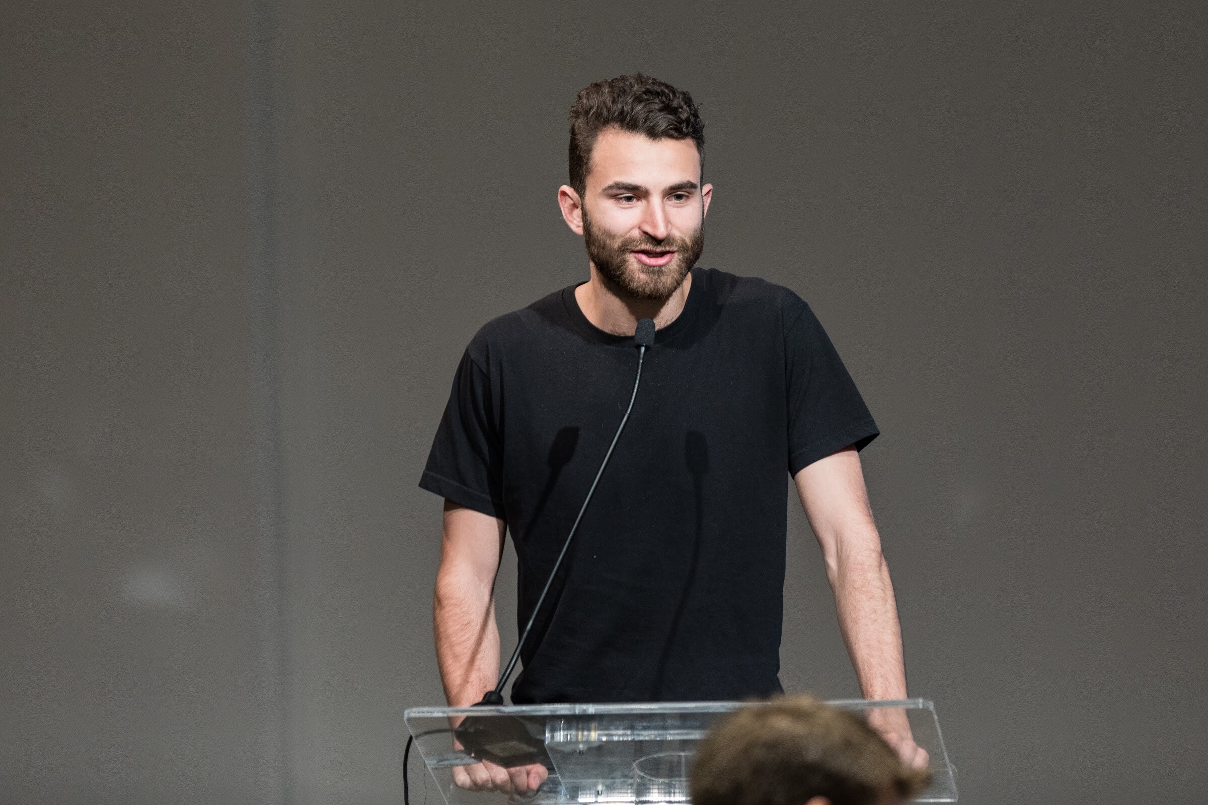 A bearded man in a black t-shirt stands behind a clear lectern with a gooseneck microphone, looking slightly down at the audience.