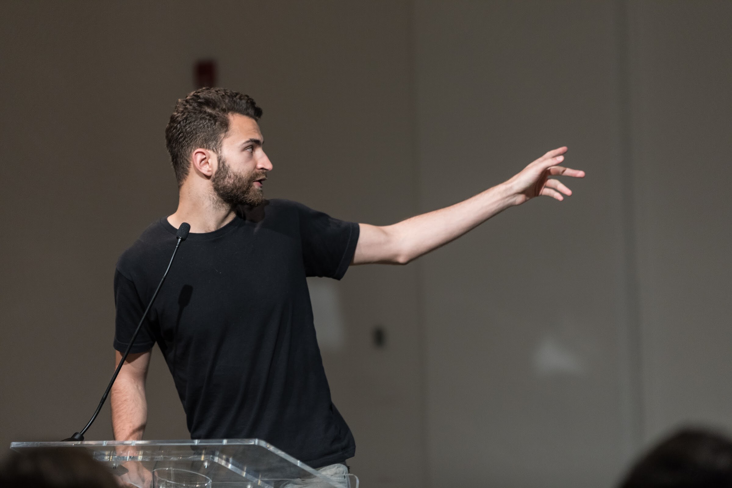 A man in a black t-shirt reaches an arm out to the right behind a clear lectern.