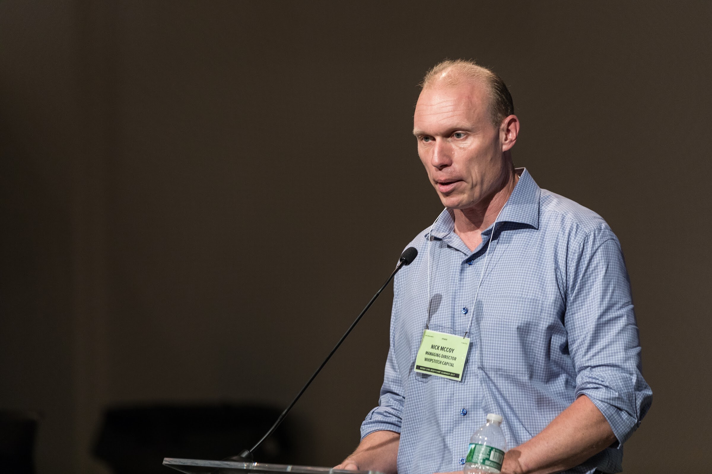 A man in a blue checked shirt with a green name badge speaks into a lectern microphone against a dark wall.