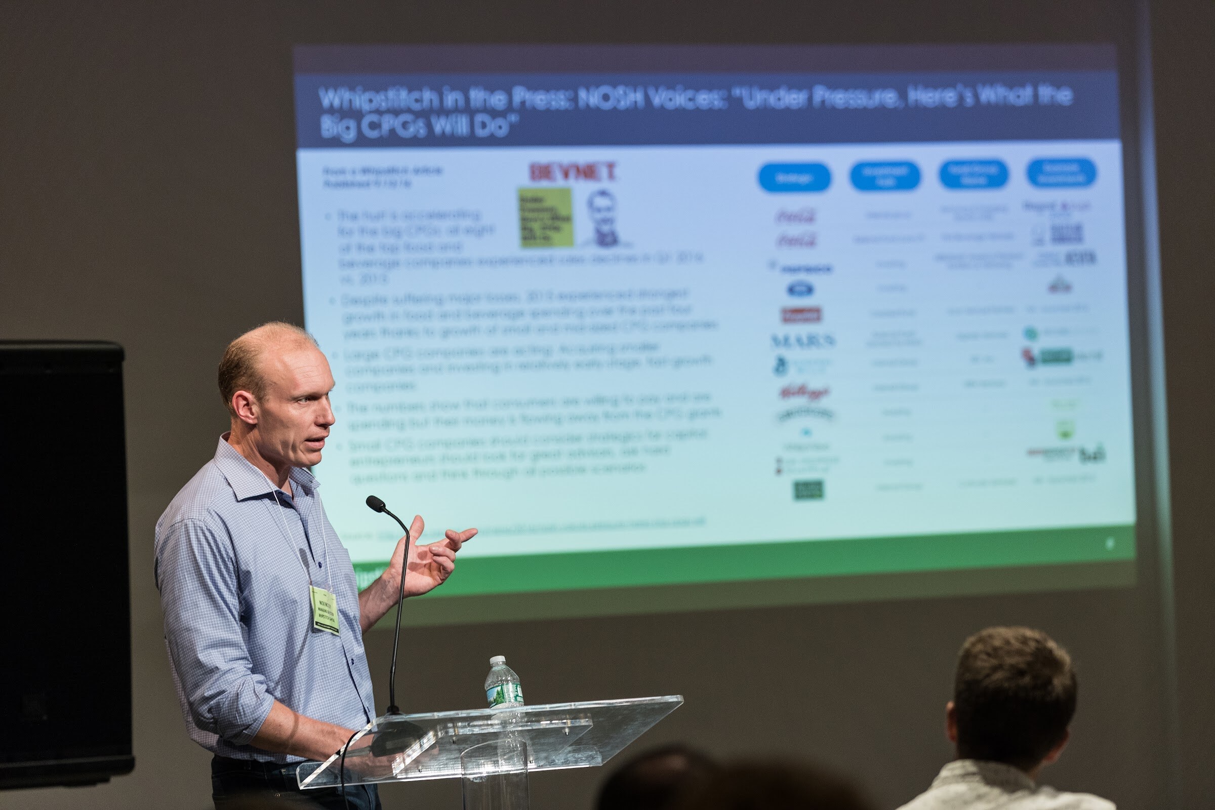 A man in a blue shirt gestures beside a clear lectern while a projected slide titled Whipstitch in the Press shows logos of large food companies.