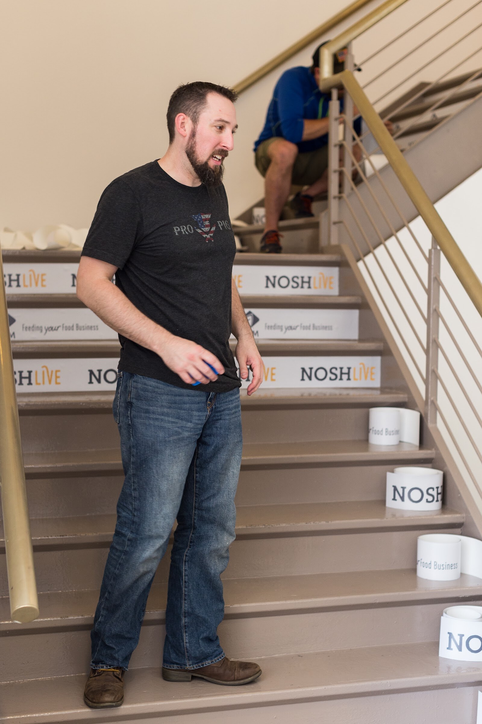 A bearded man in a gray t-shirt stands on a stairway whose risers carry Nosh Live and ADM decals, while a second worker crouches higher up.