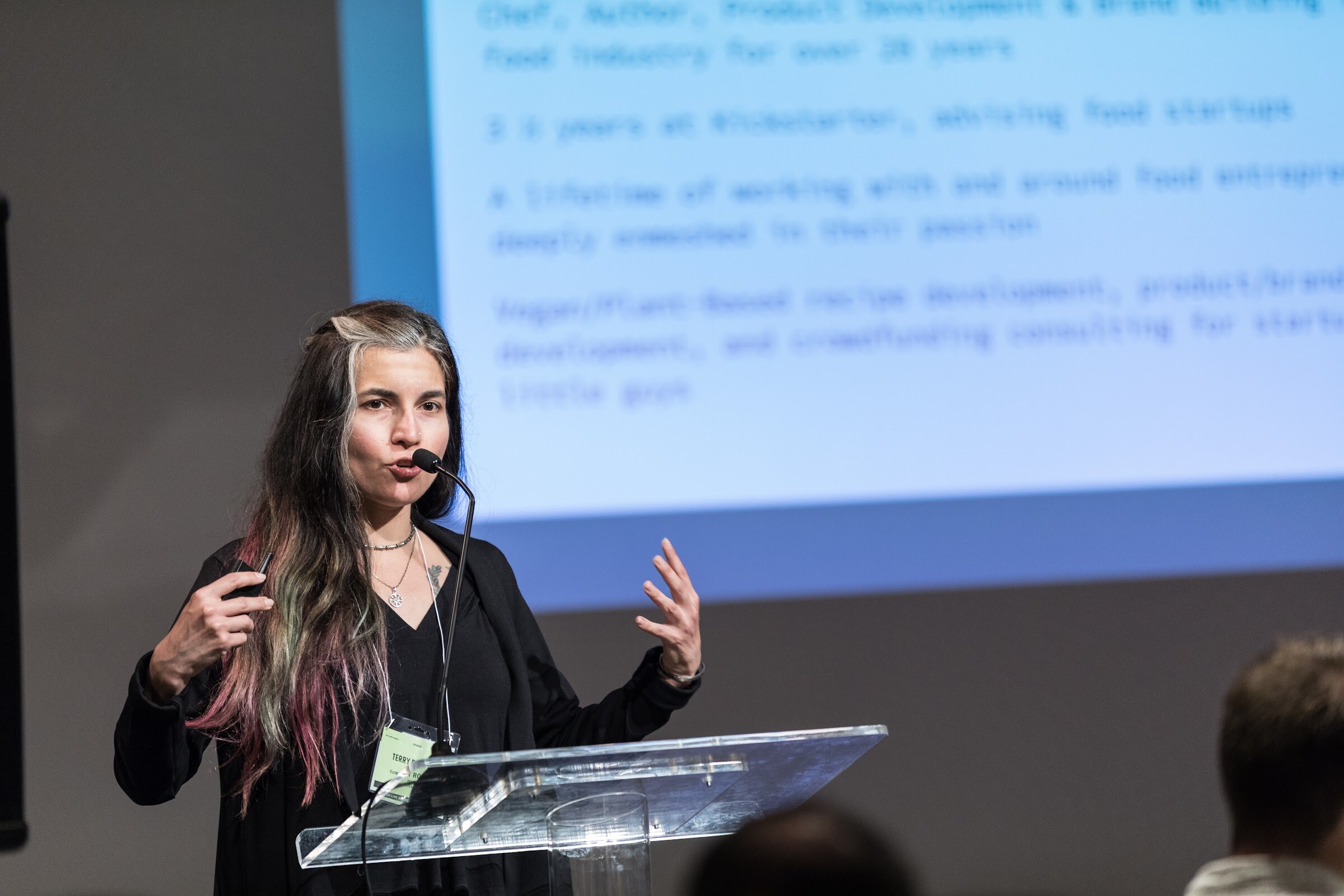 A woman with long hair in a black jacket speaks into a headset microphone behind a clear lectern, with a projected biography slide above her.