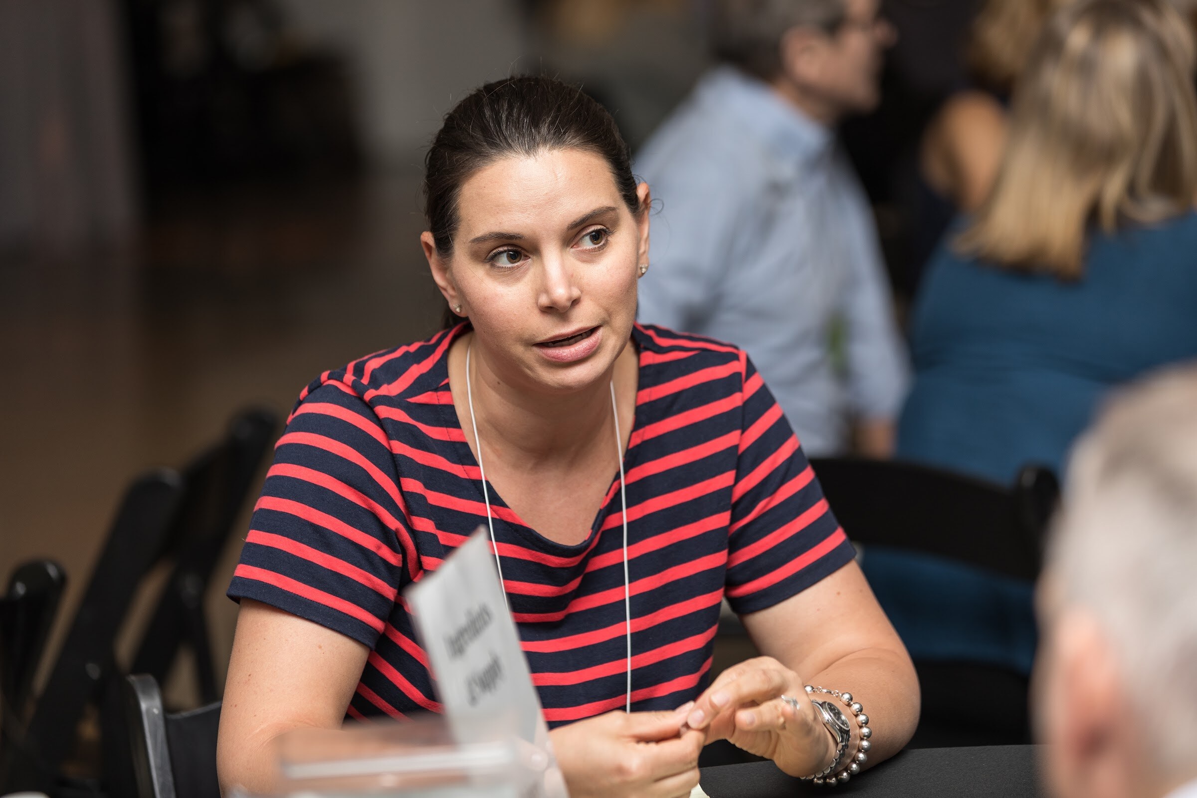 A woman in a navy and red striped top gestures while speaking at a black-clothed table, with a small sign beside her.