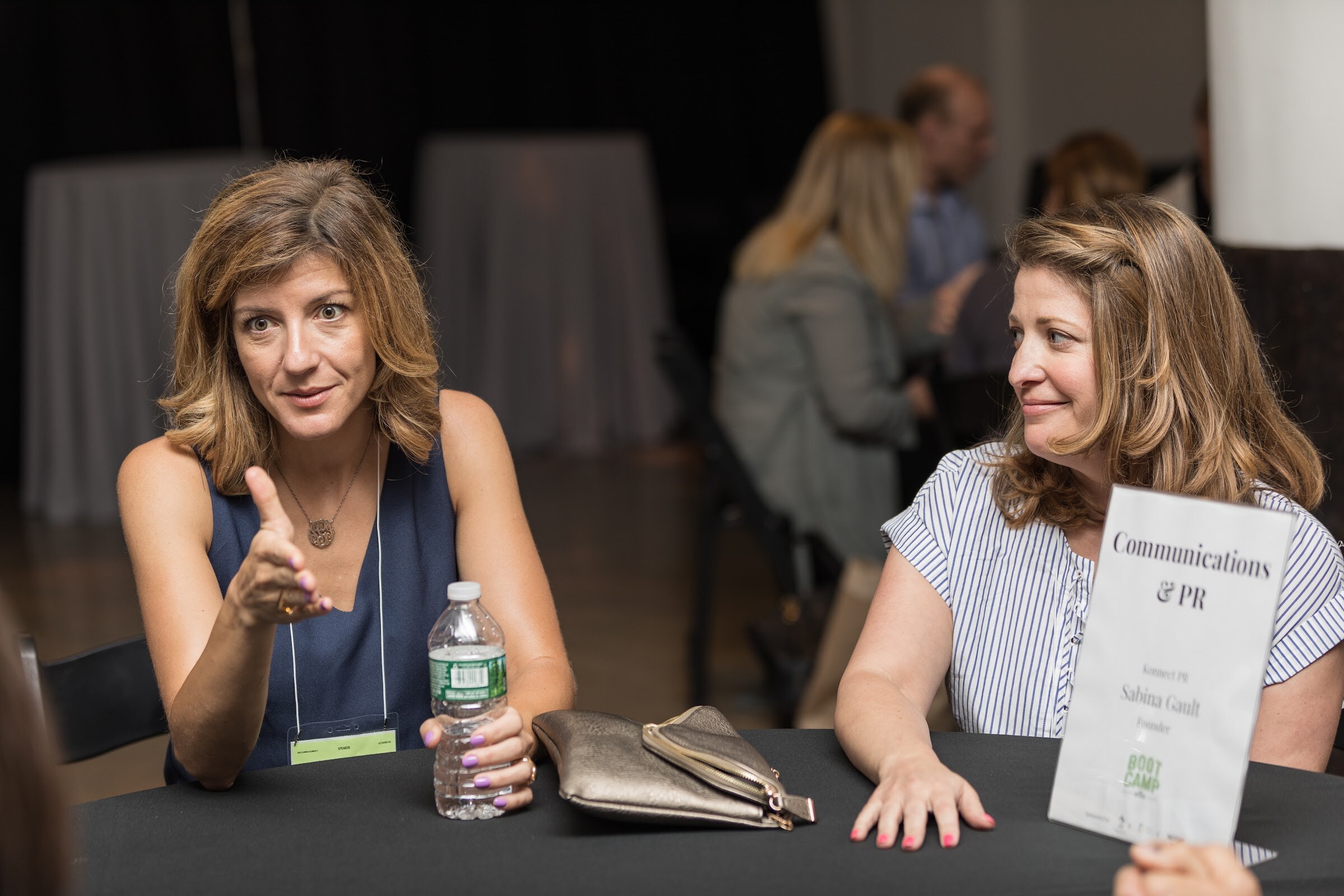 Two women sit at a black-covered table; one gestures while speaking, and a card reading Communications and PR stands at the right.