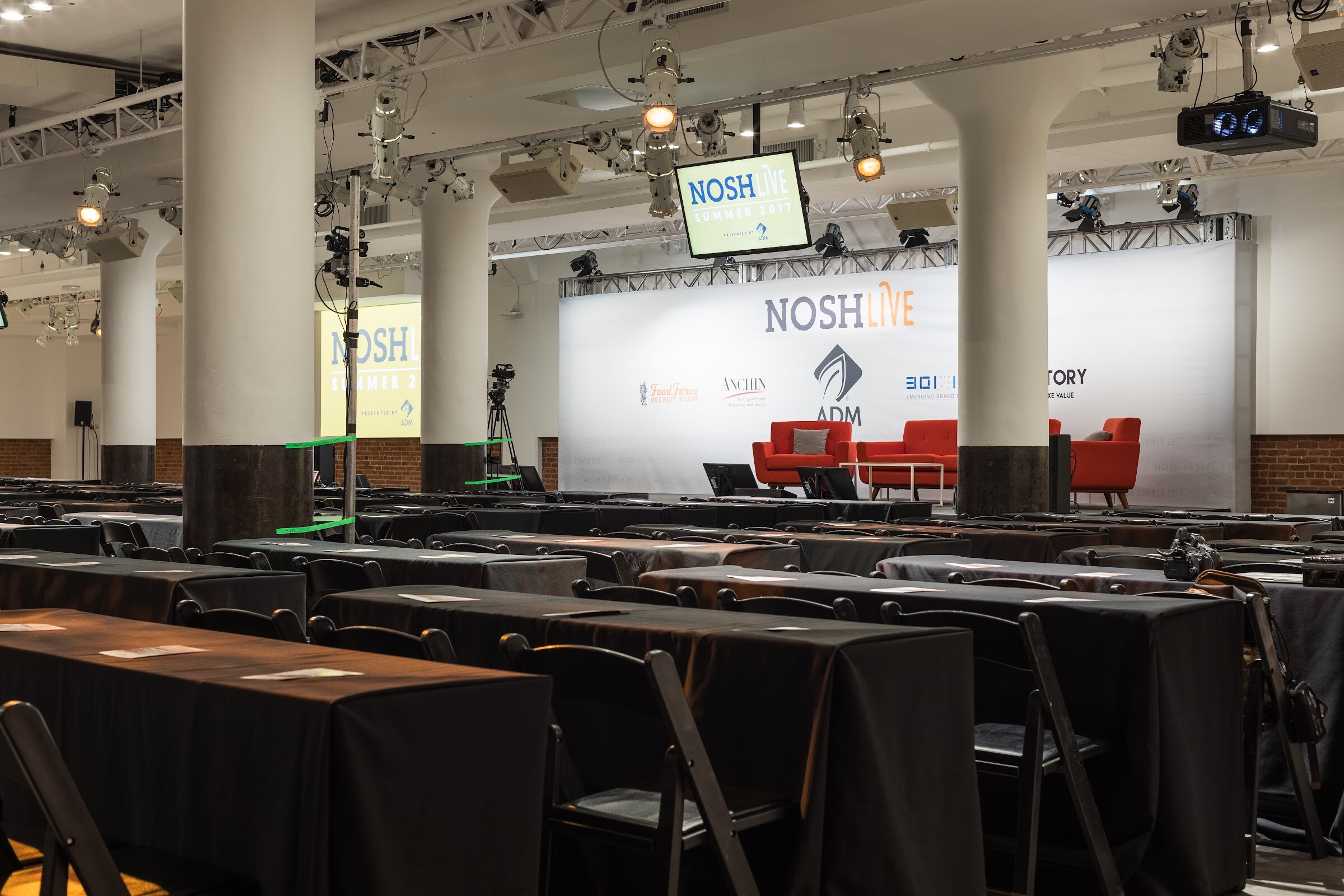 A wide room of columns, spotlights and rows of black-clothed tables facing a stage with a white Nosh Live backdrop and orange sofas.