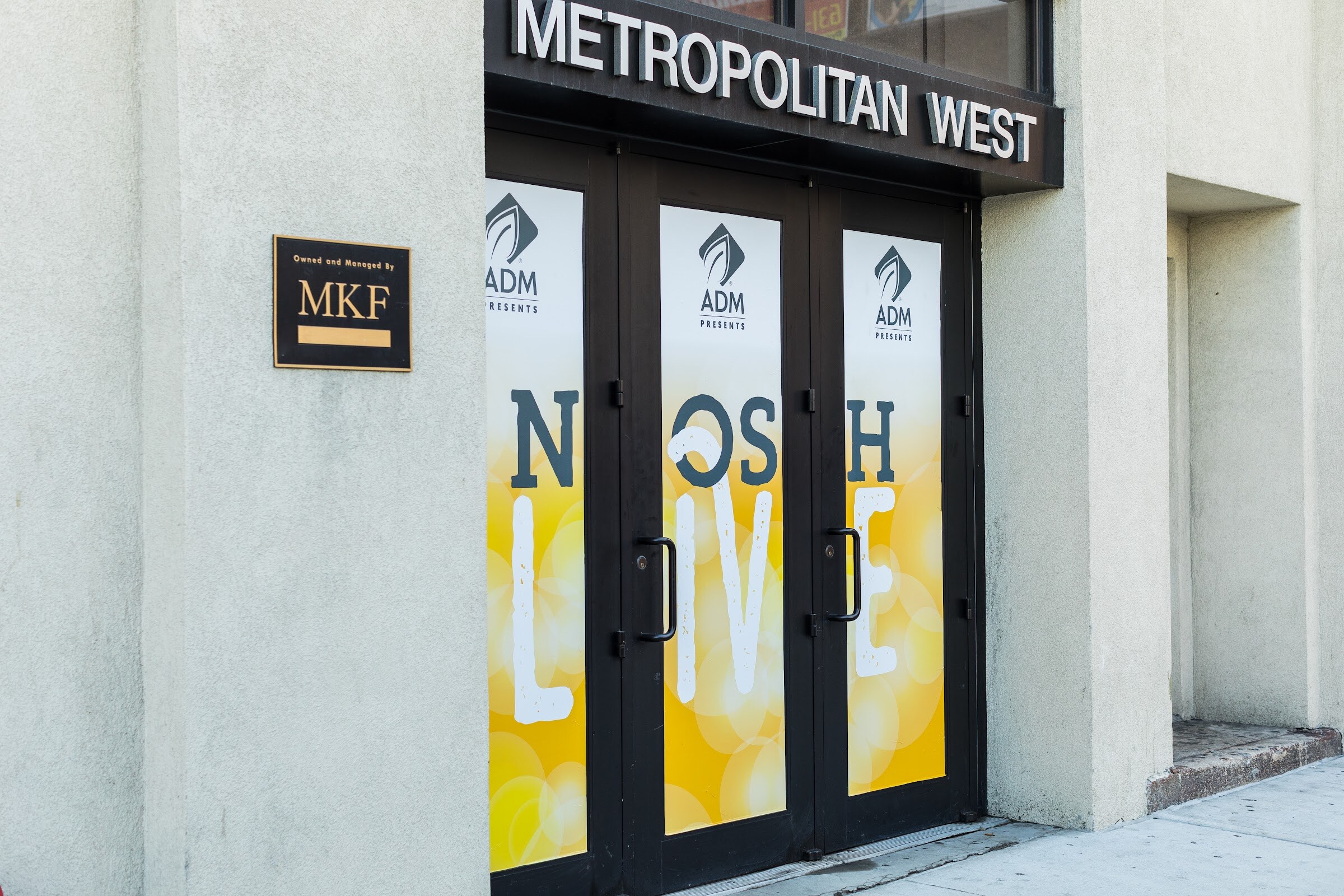 Black double doors under a Metropolitan West sign are covered in yellow decals reading ADM Presents Nosh Live.