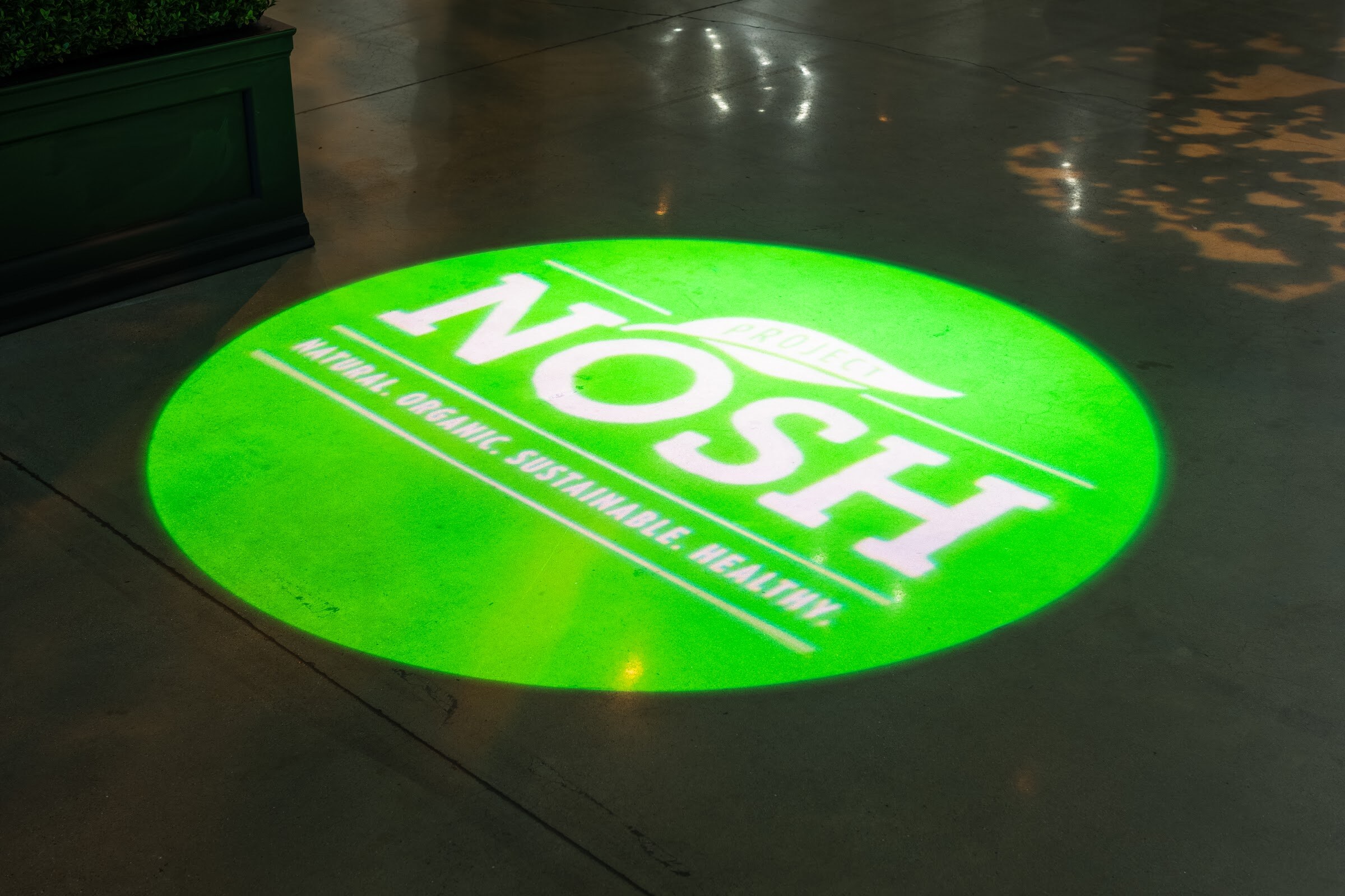 A circular green light projection on a floor shows the words Project Nosh, Natural. Organic. Sustainable. Healthy.