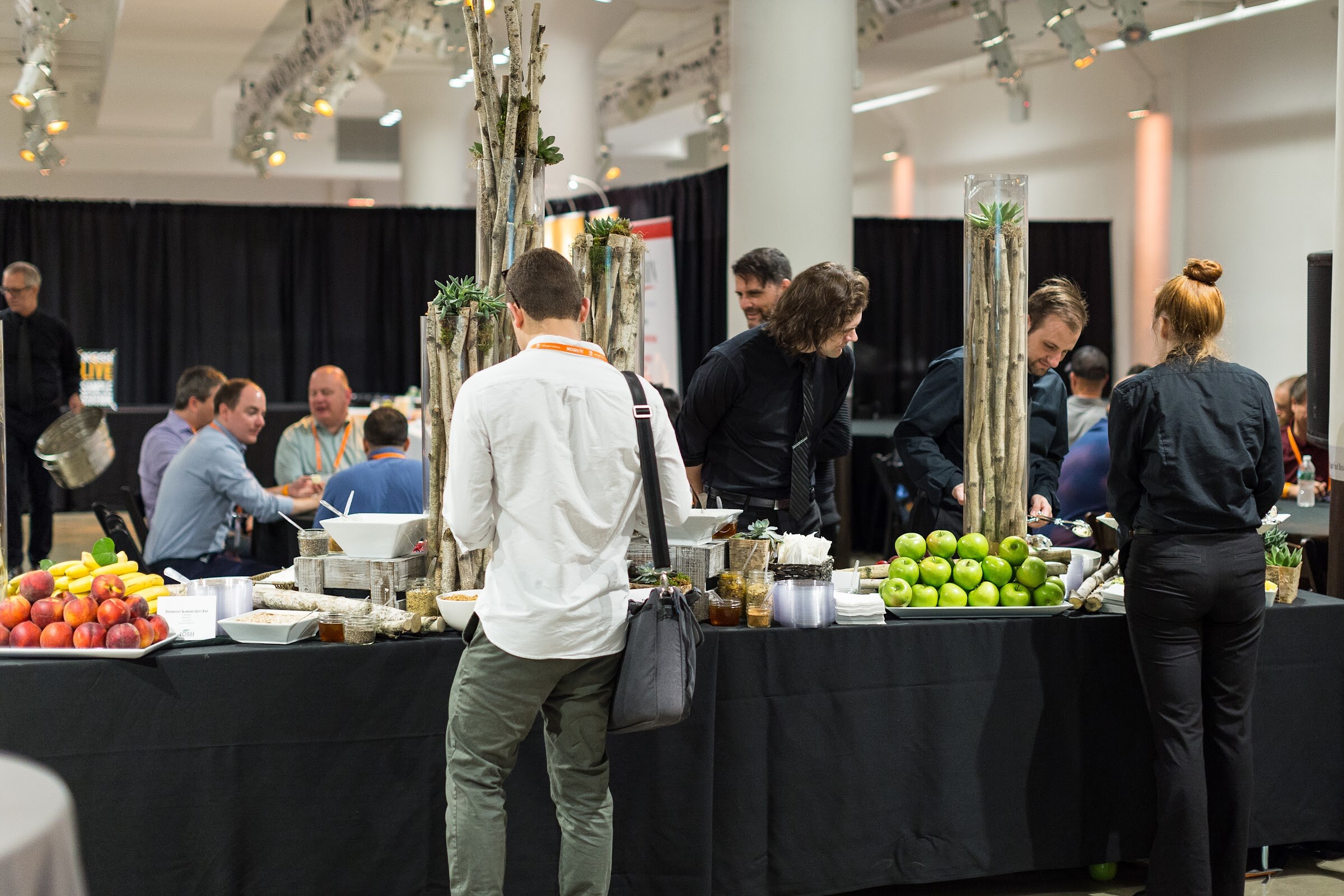 A catering buffet with fruit and succulents, with staff in black and attendees in orange lanyards serving themselves.