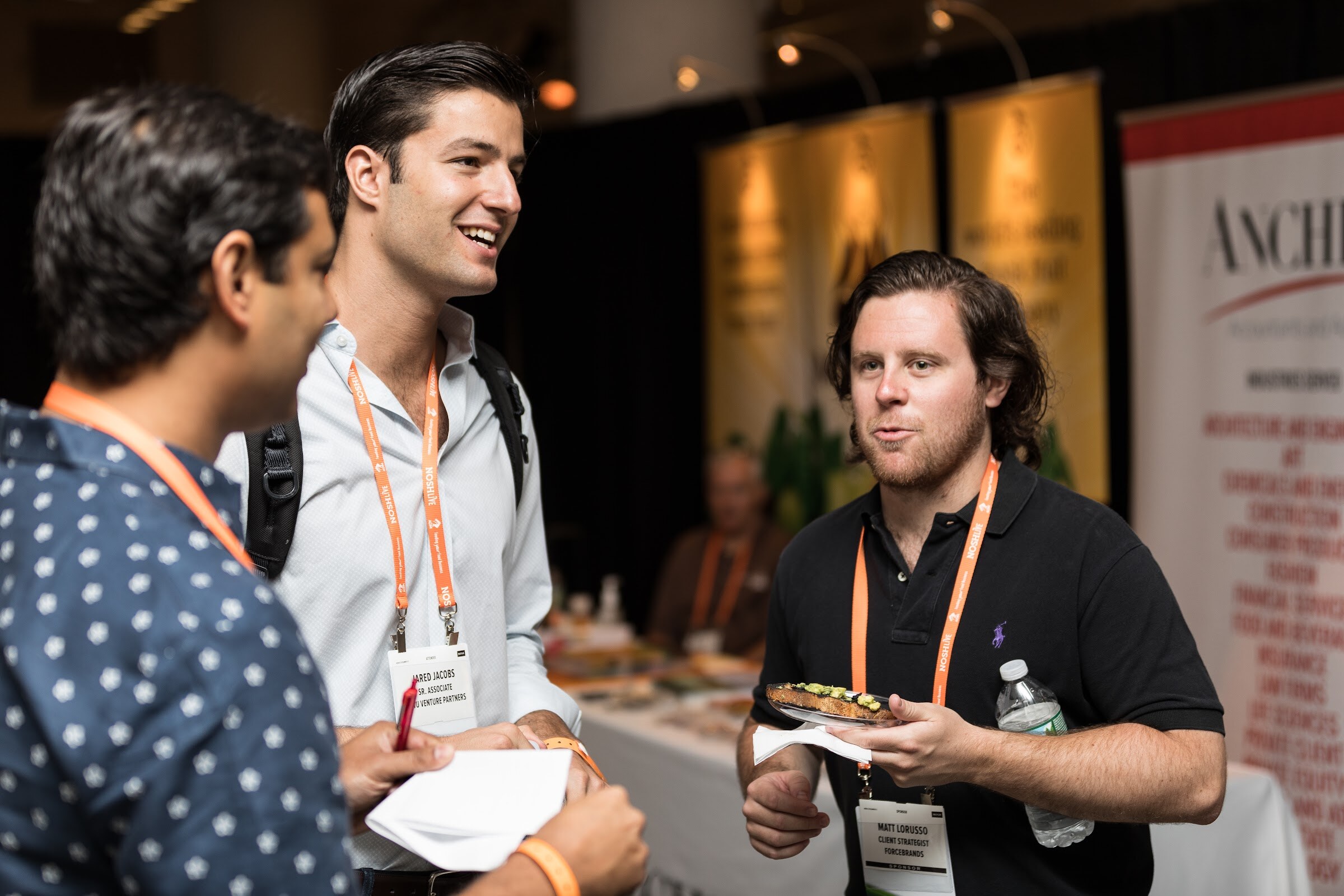 Three men in orange lanyards chat and smile, one holding a small plate, in front of sponsor tables and an Anchin banner.