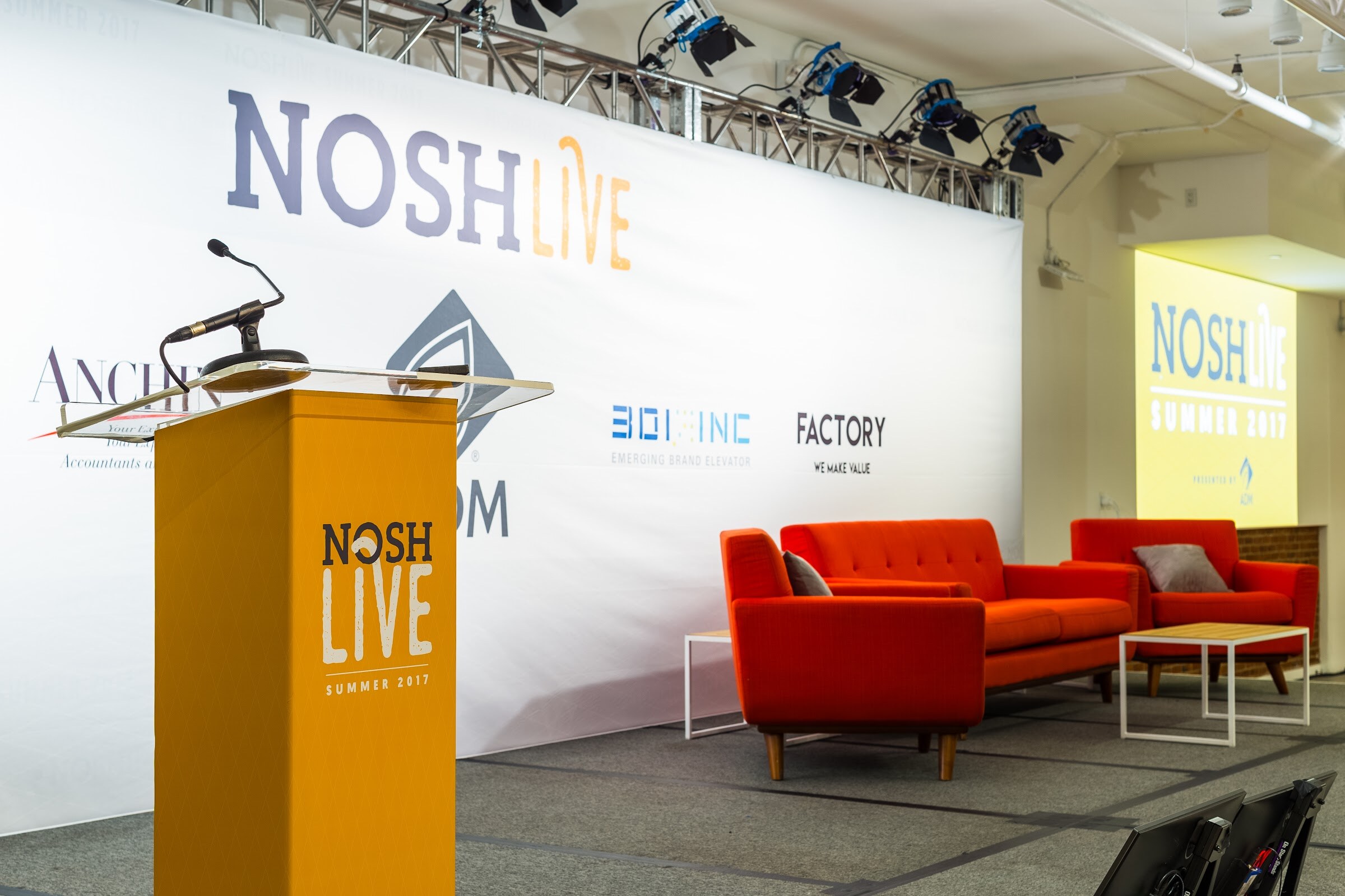A yellow Nosh Live Summer 2017 podium beside orange sofas on a stage with a backdrop showing ADM, Anchin, 301 Inc and Factory.