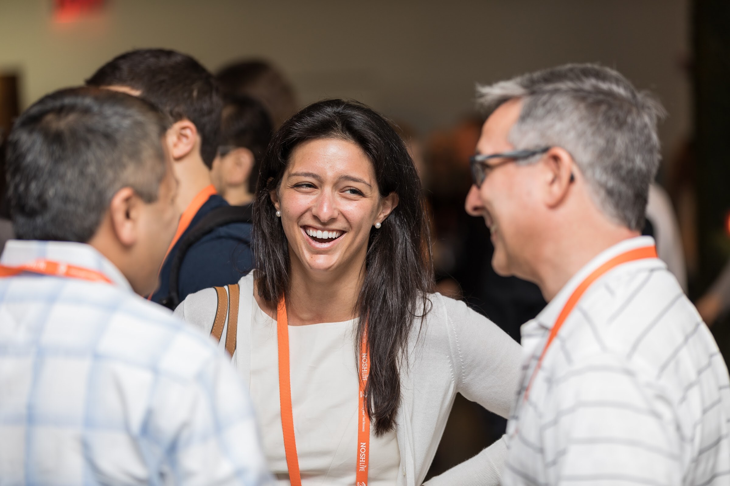 A smiling woman in a white cardigan with an orange Nosh Live lanyard talks with two men in the foreground.