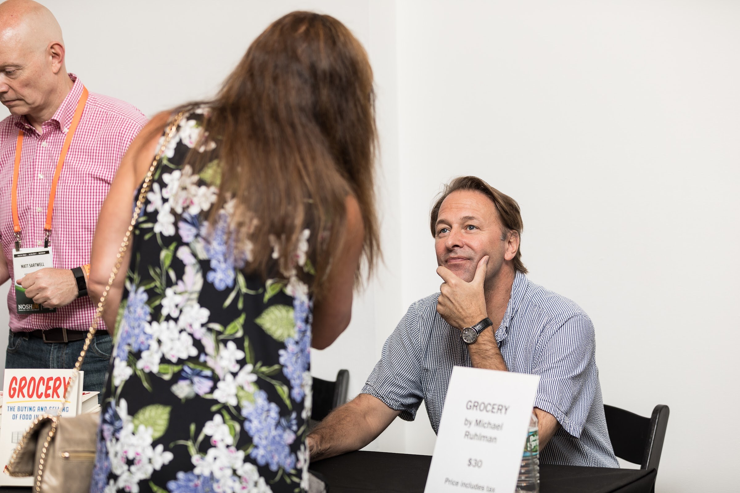 A man in a striped shirt sits at a table with a Grocery book sign and listens to a woman in a floral dress standing in front of him.