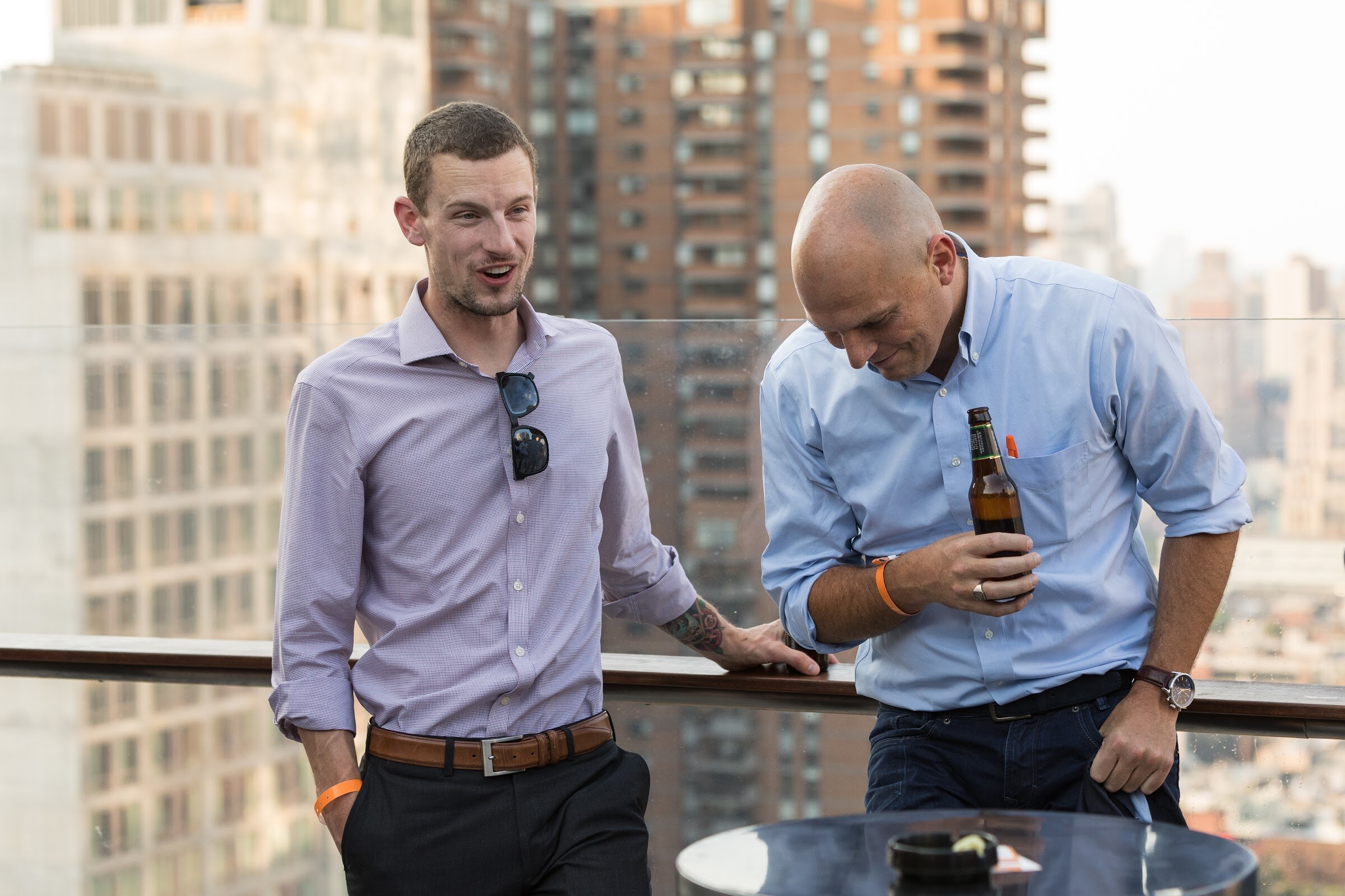 Two men lean on a glass railing in front of apartment towers, one holding a beer bottle.