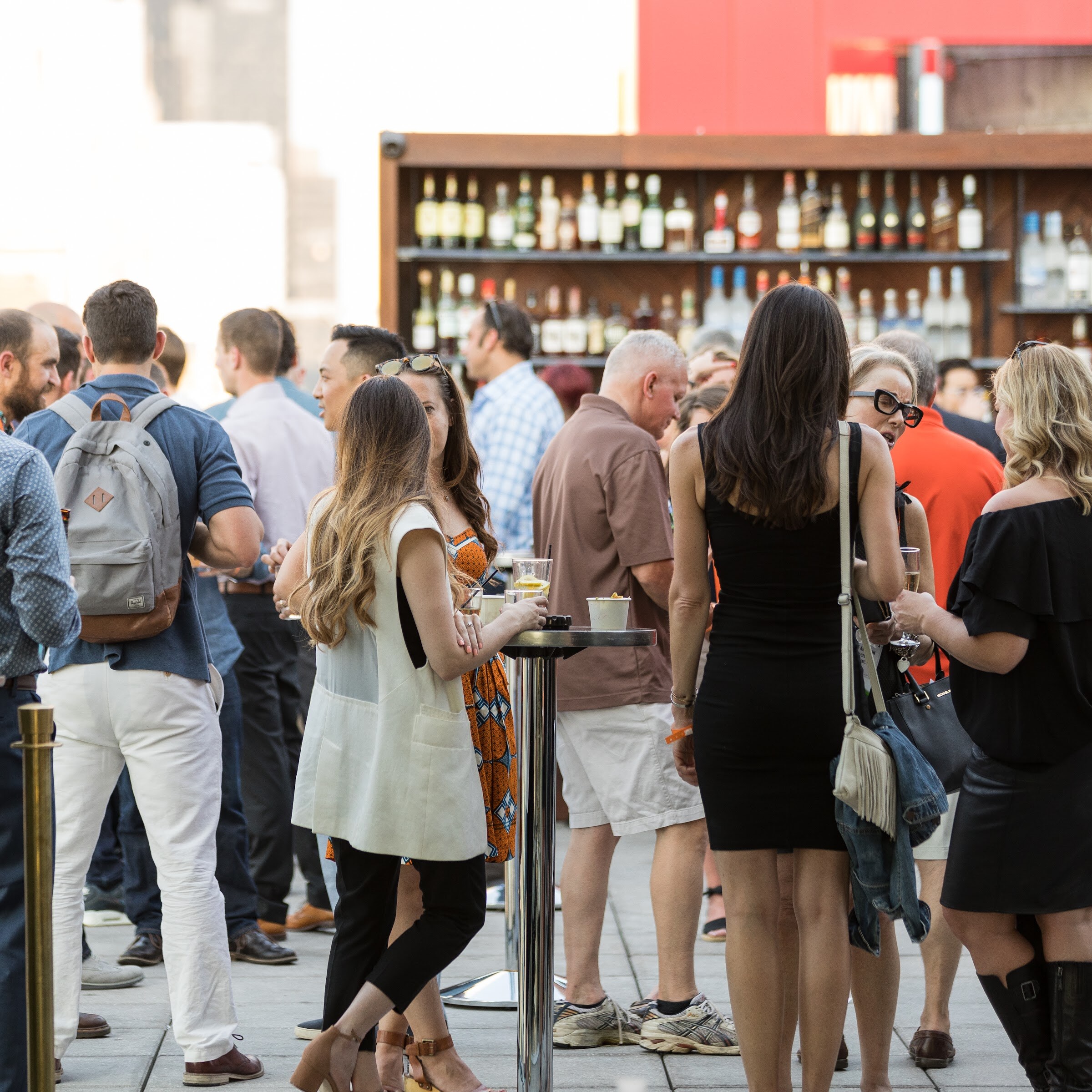 A crowd of people stands on a rooftop terrace talking and holding drinks in front of a wooden bar with shelves of liquor bottles.