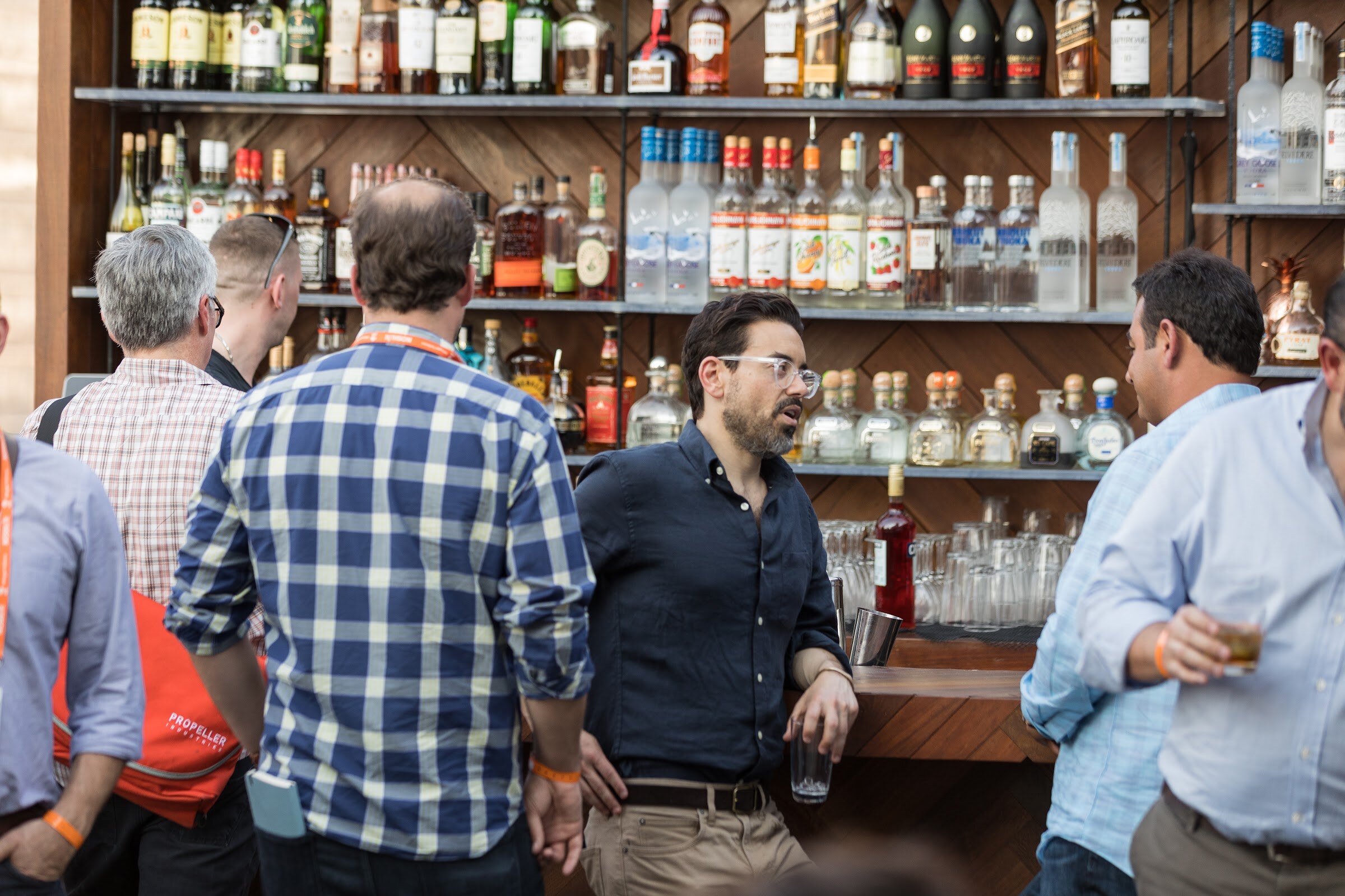 A bearded man in a navy shirt talks at a rooftop bar in front of shelves of liquor bottles, with other guests in the foreground.