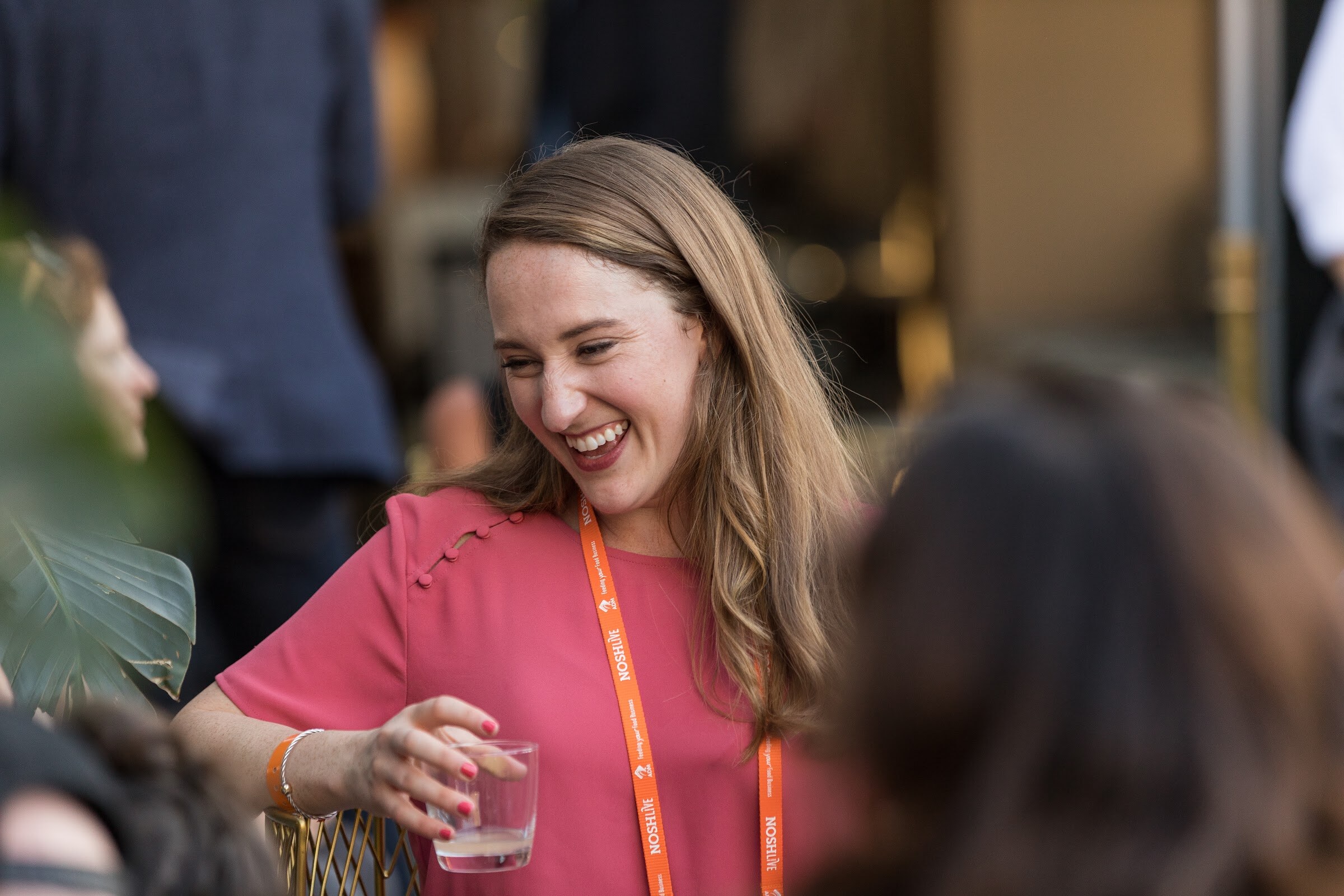 A smiling woman in a pink top and orange Nosh Live lanyard holds a glass, with a plant in the blurred foreground.