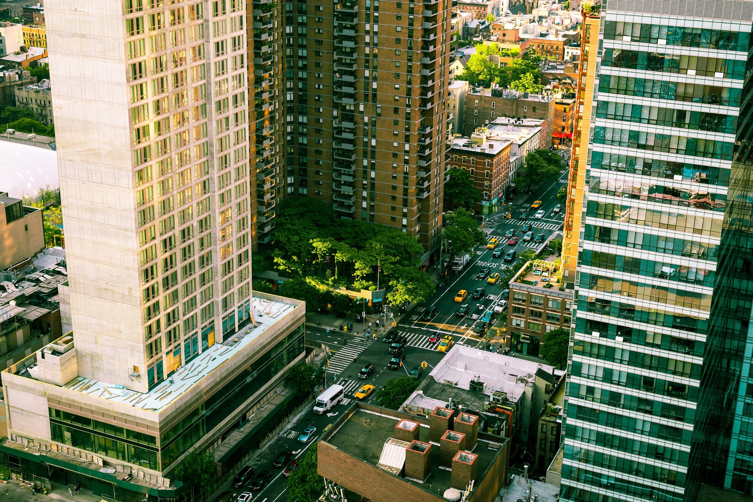 An aerial view of a city street with yellow taxis between tall brick and glass buildings and green trees.
