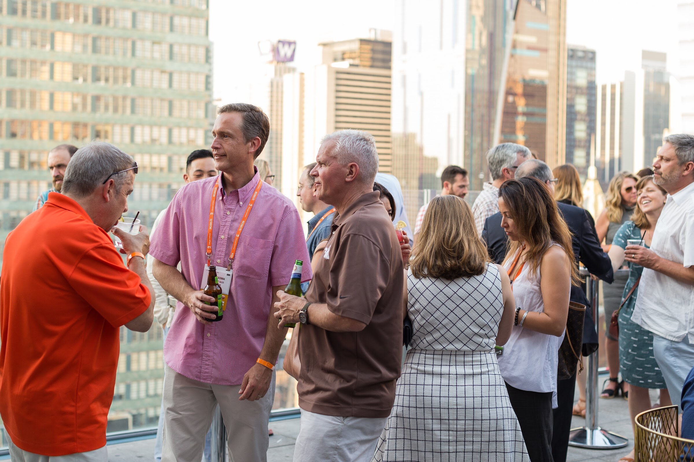 A group of attendees with orange bracelets and drinks stand on a rooftop terrace with high-rise buildings behind them.