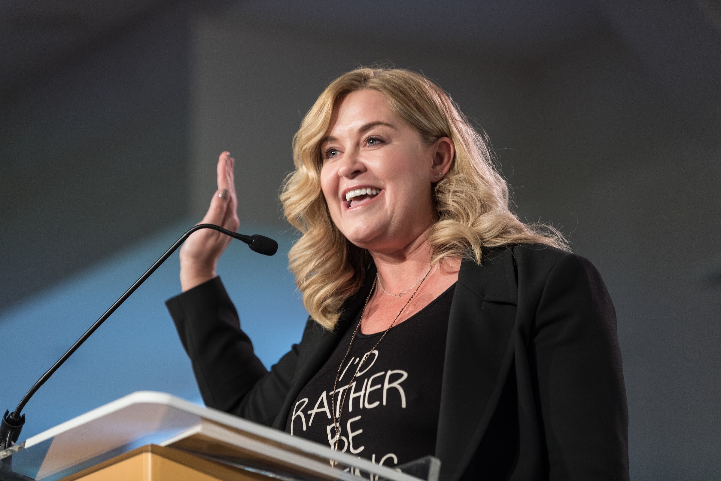 A smiling woman with wavy blonde hair in a black blazer and graphic t-shirt speaks at a podium with her hand raised.