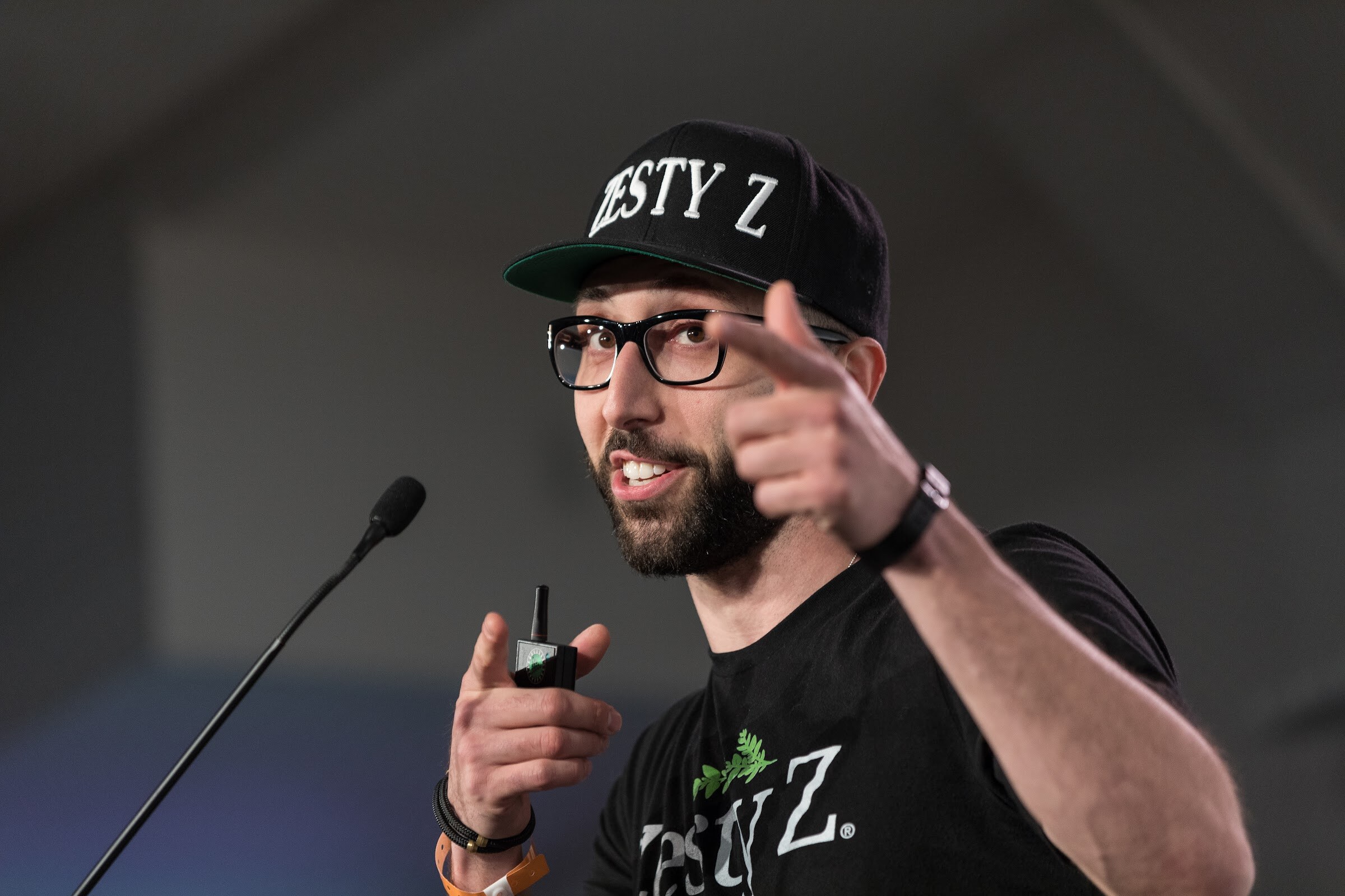 A bearded man in a black Zesty Z cap and t-shirt points with one hand and holds a clicker with the other beside a microphone.