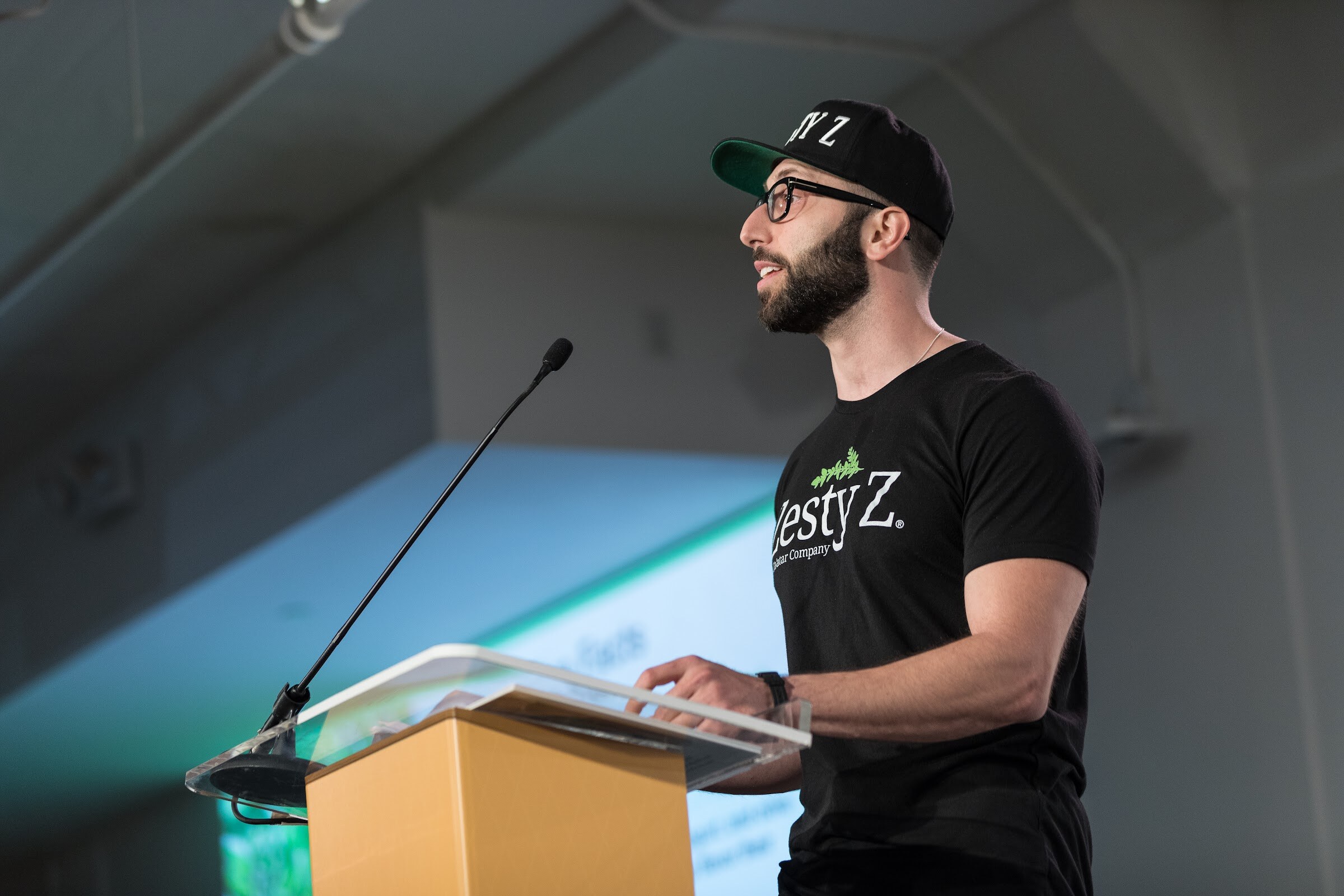 A bearded man in a black Zesty Z cap and shirt stands at a clear podium with a gooseneck microphone, a green slide projected behind.