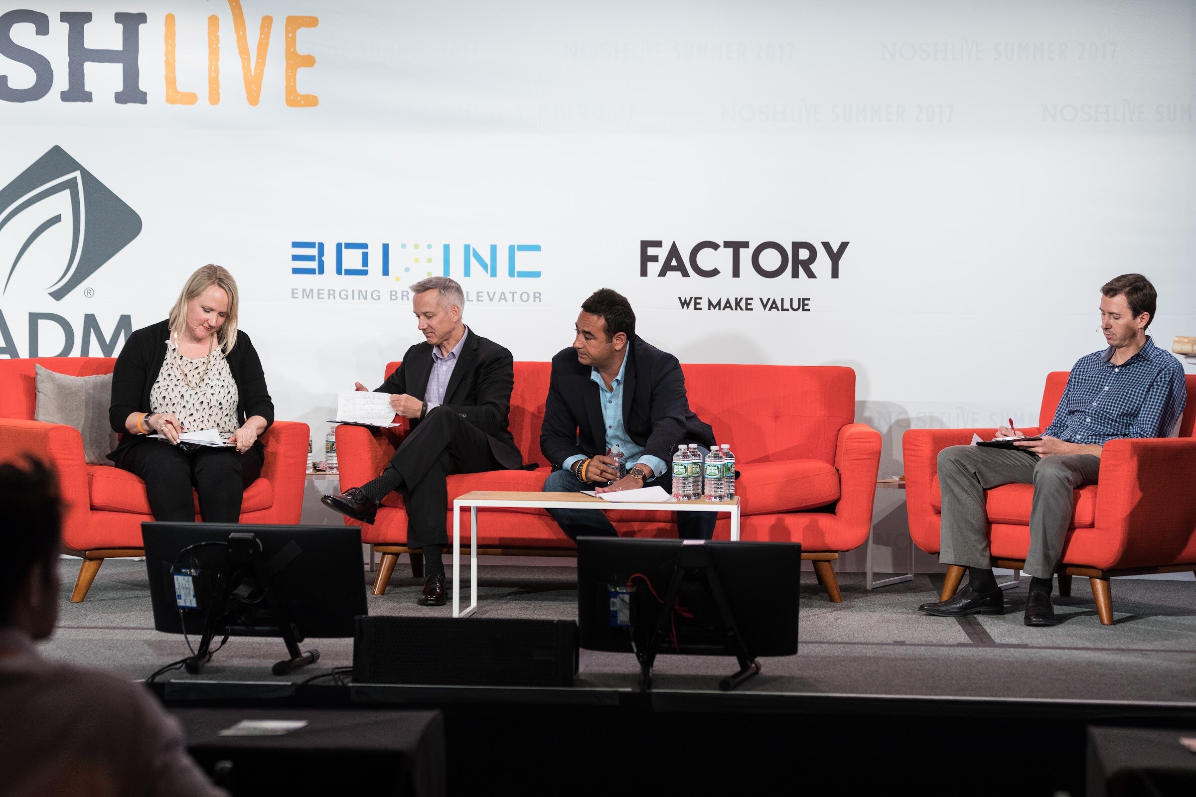 Four people sit on orange sofas and armchairs on stage in front of 301 Inc and Factory banners, two looking at papers and one writing in a notebook.