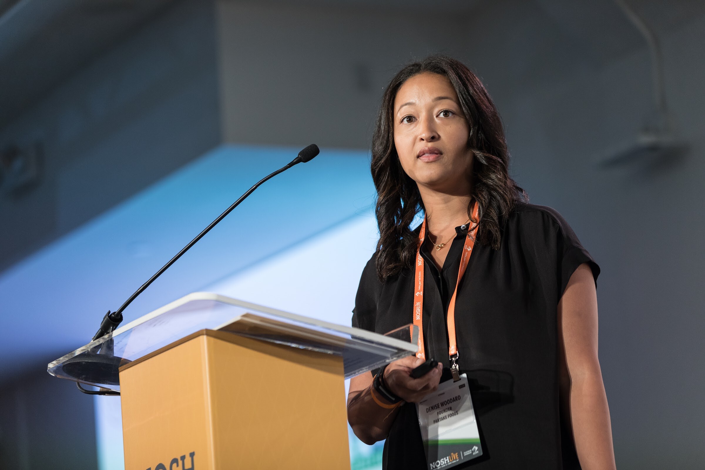 A woman with long dark hair and an orange lanyard stands at a Nosh Live podium; her badge reads Partake Foods.