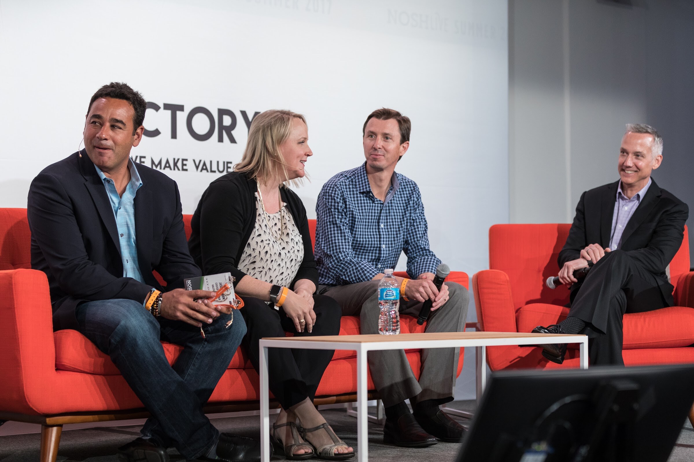 Four smiling people sit on orange sofas, one holding a microphone and another holding a lanyard, with a Factory banner behind them.
