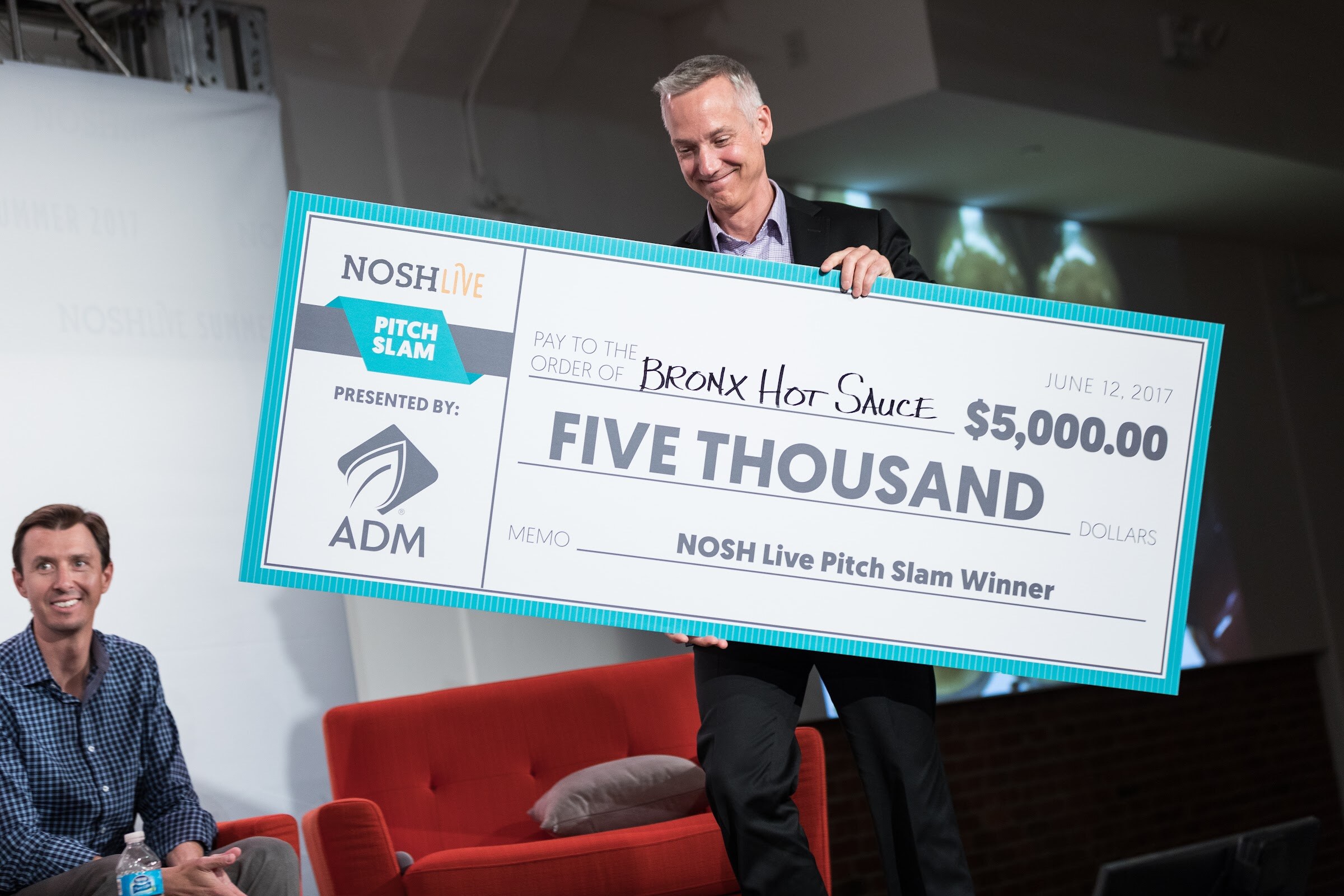 A man in a dark suit holds a large check made out to Bronx Hot Sauce for $5,000, presented by ADM, while a seated man in a blue checked shirt smiles at the lower left.