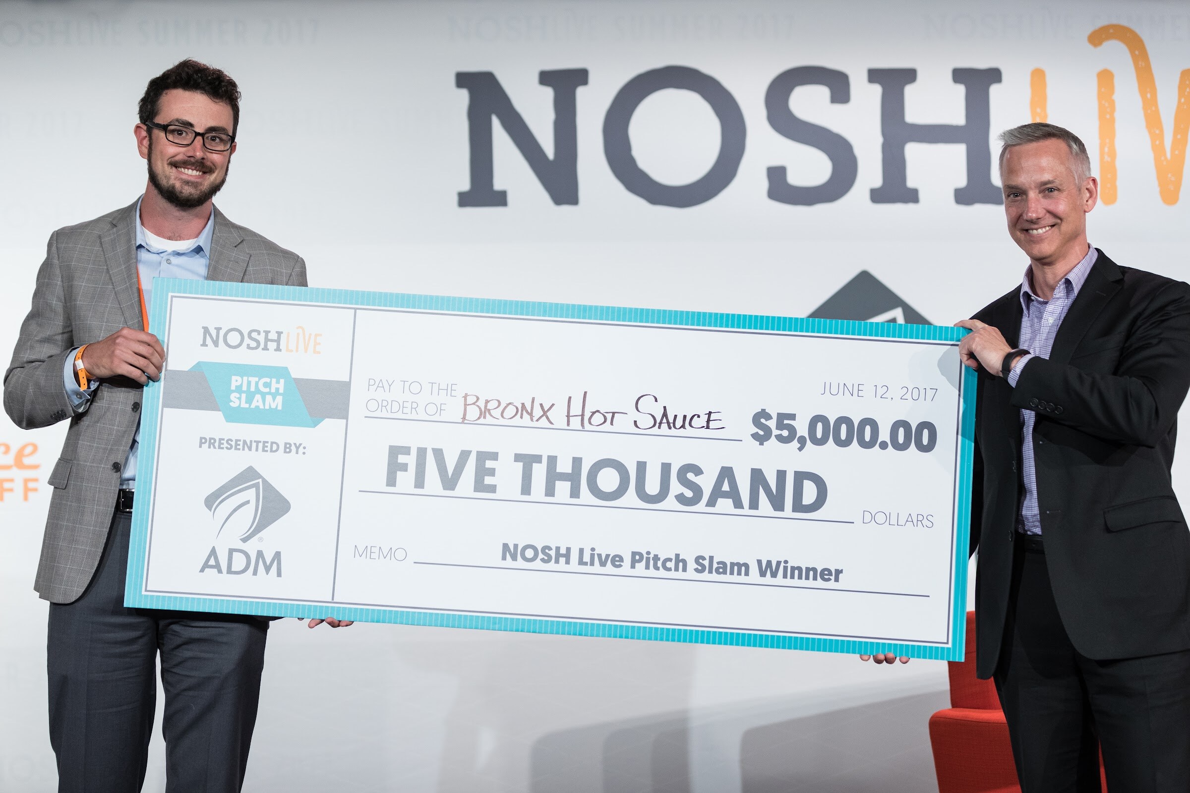 Two smiling men hold a large Nosh Live Pitch Slam check for five thousand dollars made out to Bronx Hot Sauce and dated June 12, 2017.