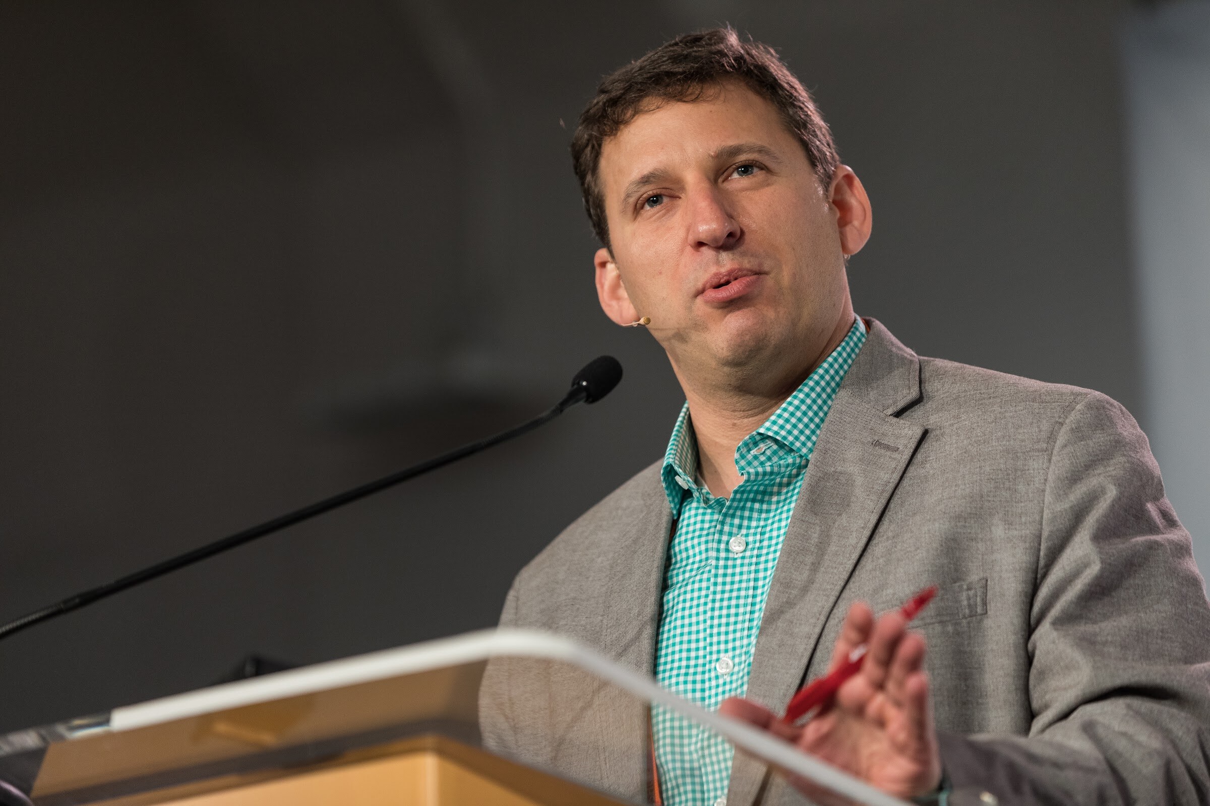 A man in a gray blazer and green gingham shirt speaks into a microphone at an orange podium, holding a red pen.