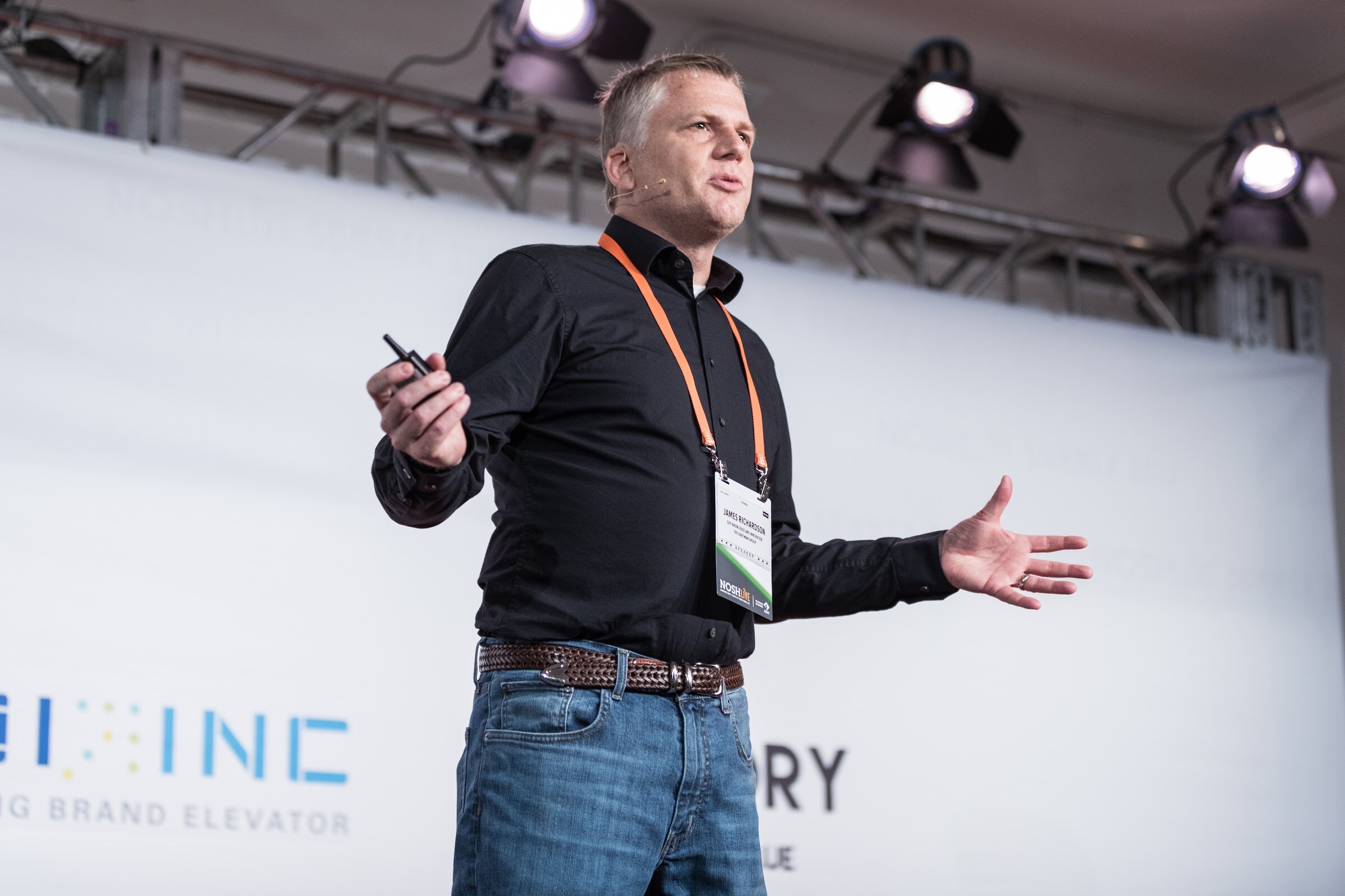 A man in a black shirt and orange lanyard stands on a stage lit by spotlights, one hand extended, in front of a white backdrop with sponsor logos.