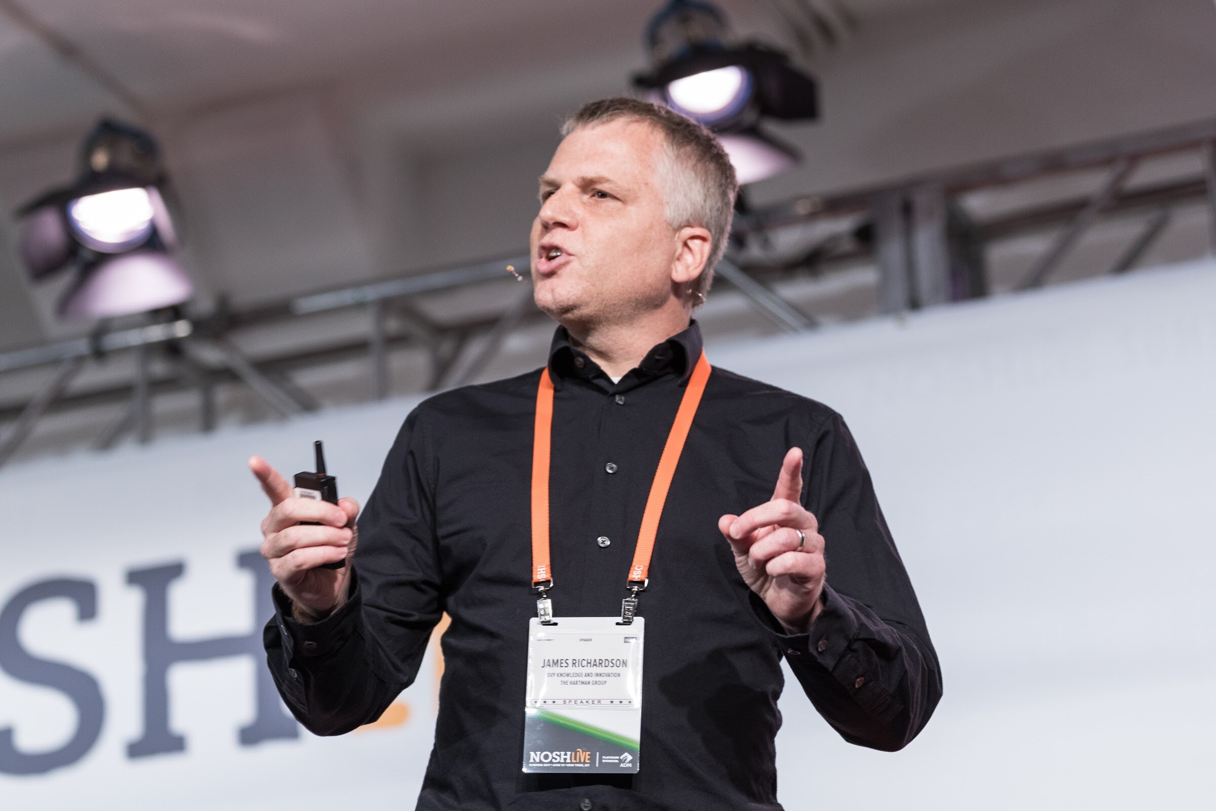 A man in a black button-up shirt with an orange lanyard and speaker badge speaks with both index fingers raised, stage lights above him.