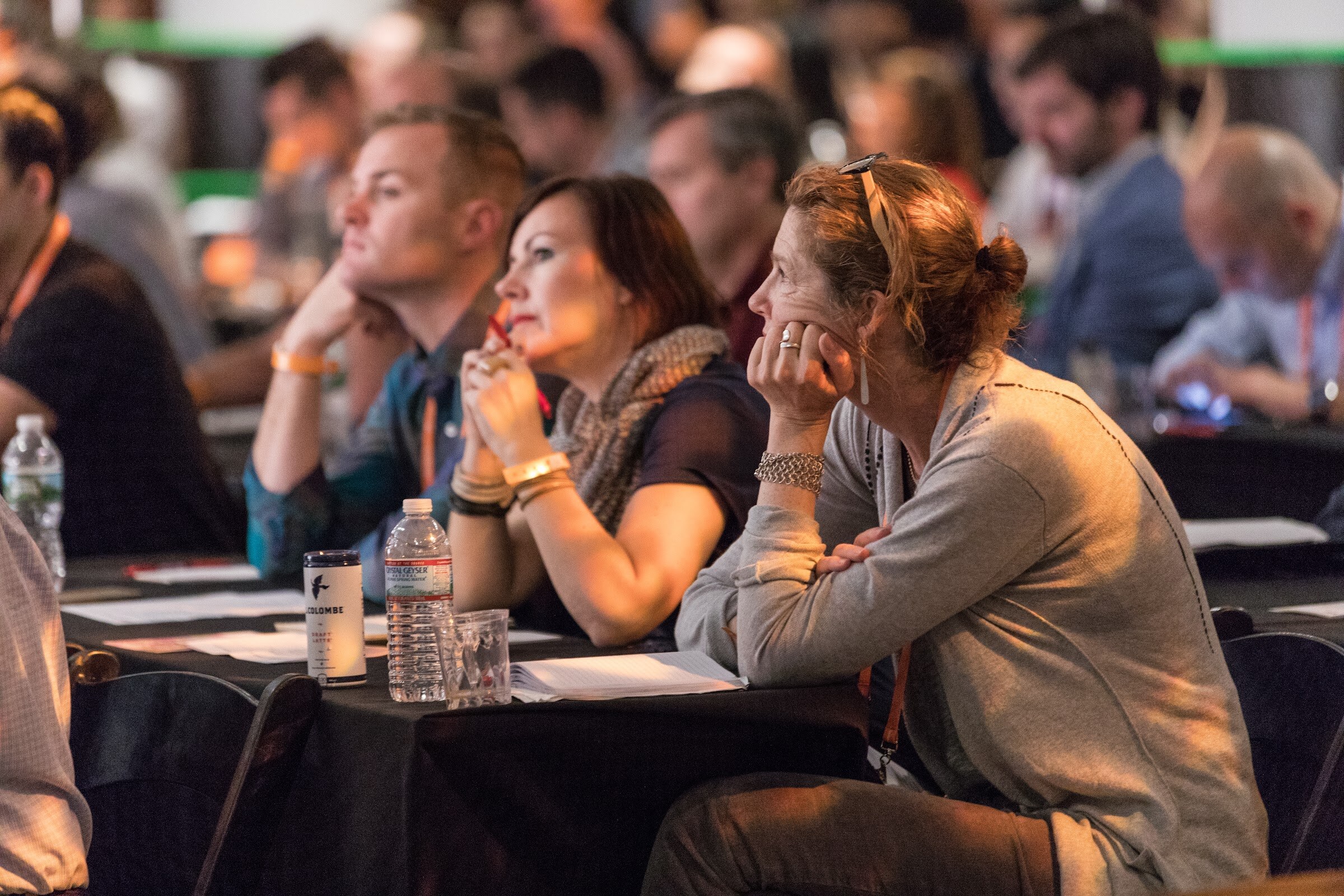 Three seated attendees with orange lanyards watch the stage intently from a black-covered table, with a water bottle and notebook in front of them.