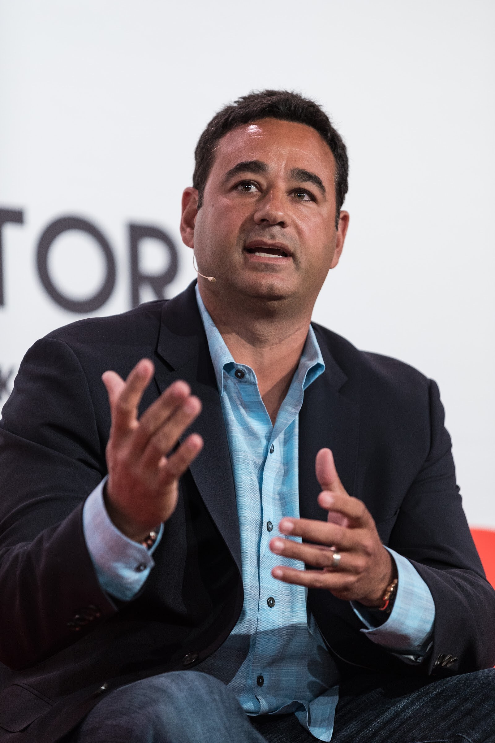 A man in a navy blazer and light blue shirt sits on an orange sofa and talks with both hands open in front of a Factory sponsor banner.