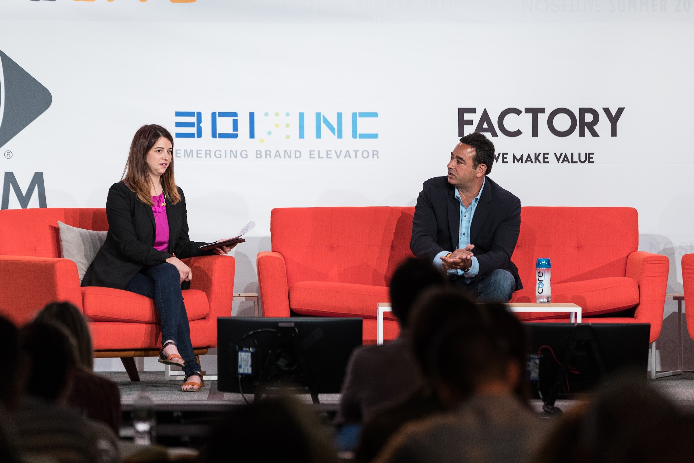 Two people sit on orange sofas on a stage in front of white sponsor banners reading 301 Inc and Factory, with audience heads in the foreground.