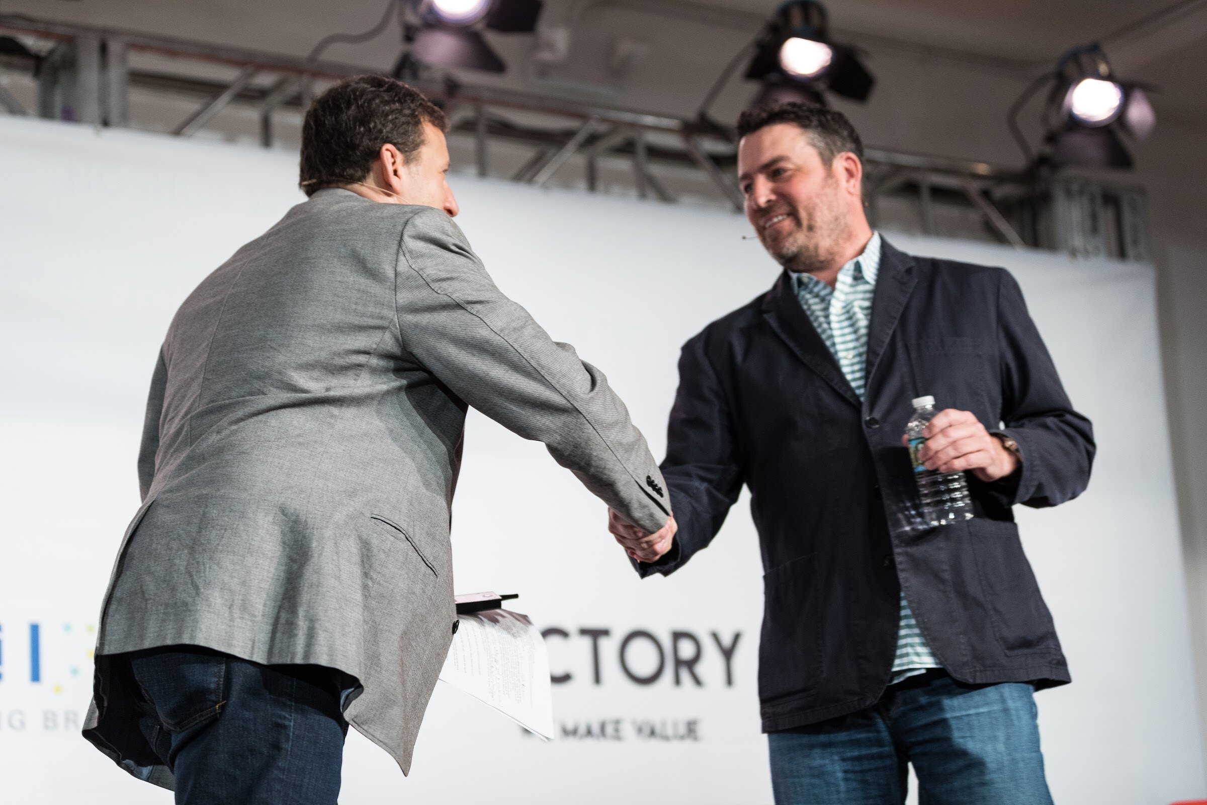 Two men in blazers shake hands on a stage beneath spotlights, one holding a water bottle.