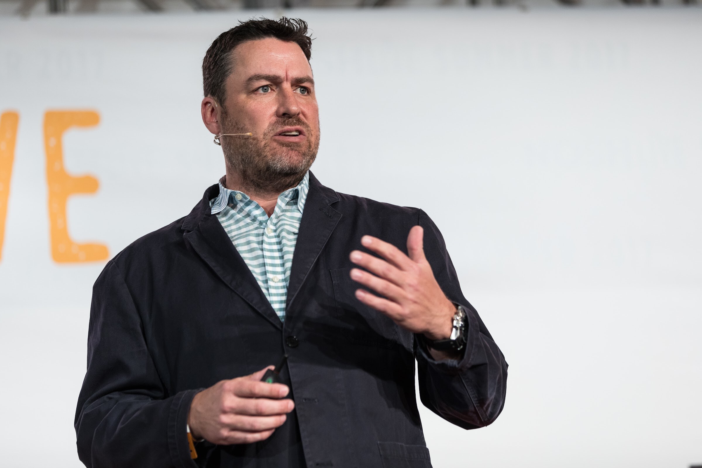 A man in a dark jacket over a striped shirt gestures with one hand and holds a clicker, with part of the orange Live logo behind him.
