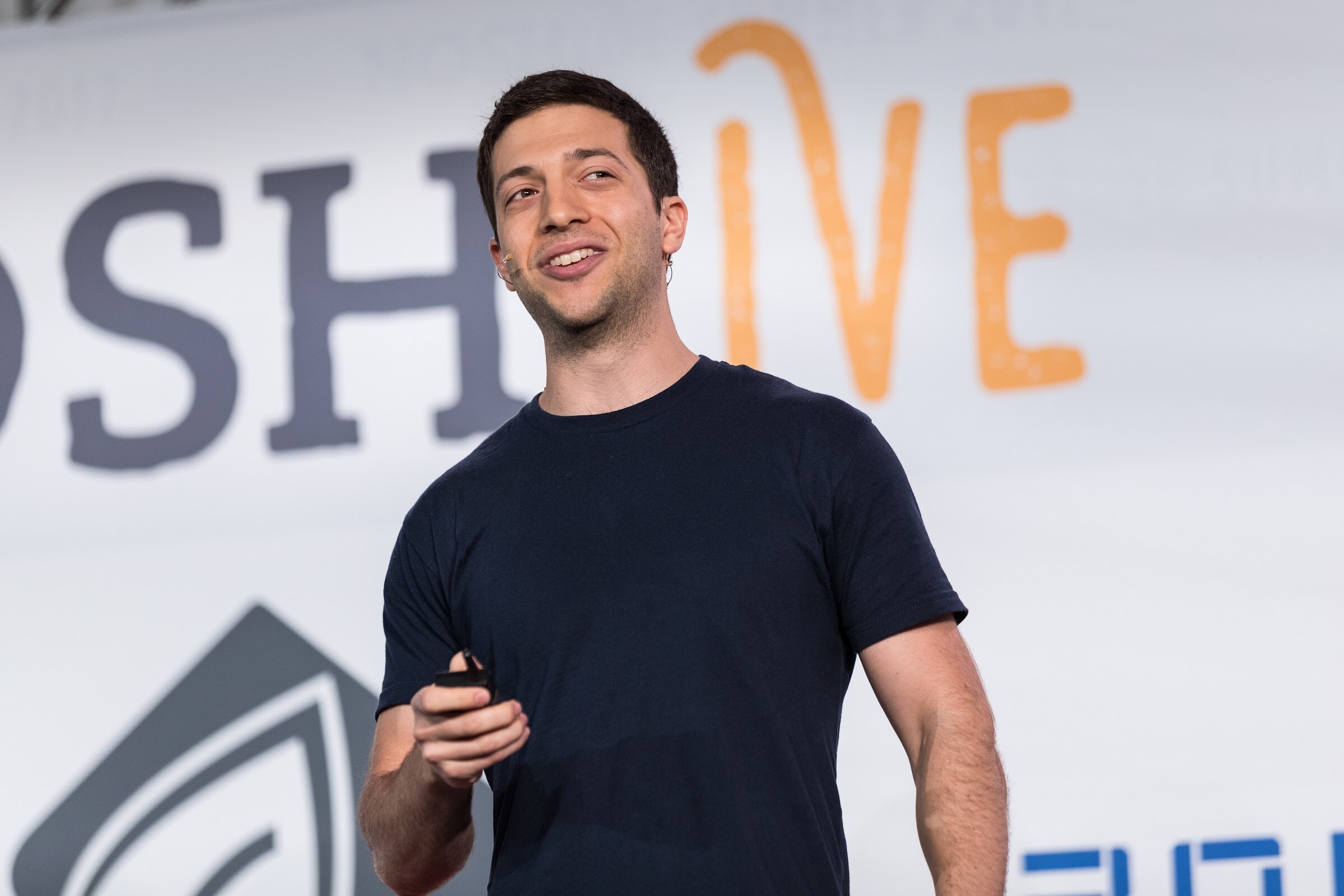 A smiling man in a dark navy t-shirt holds a presentation clicker in front of a white banner with the Nosh Live logo.