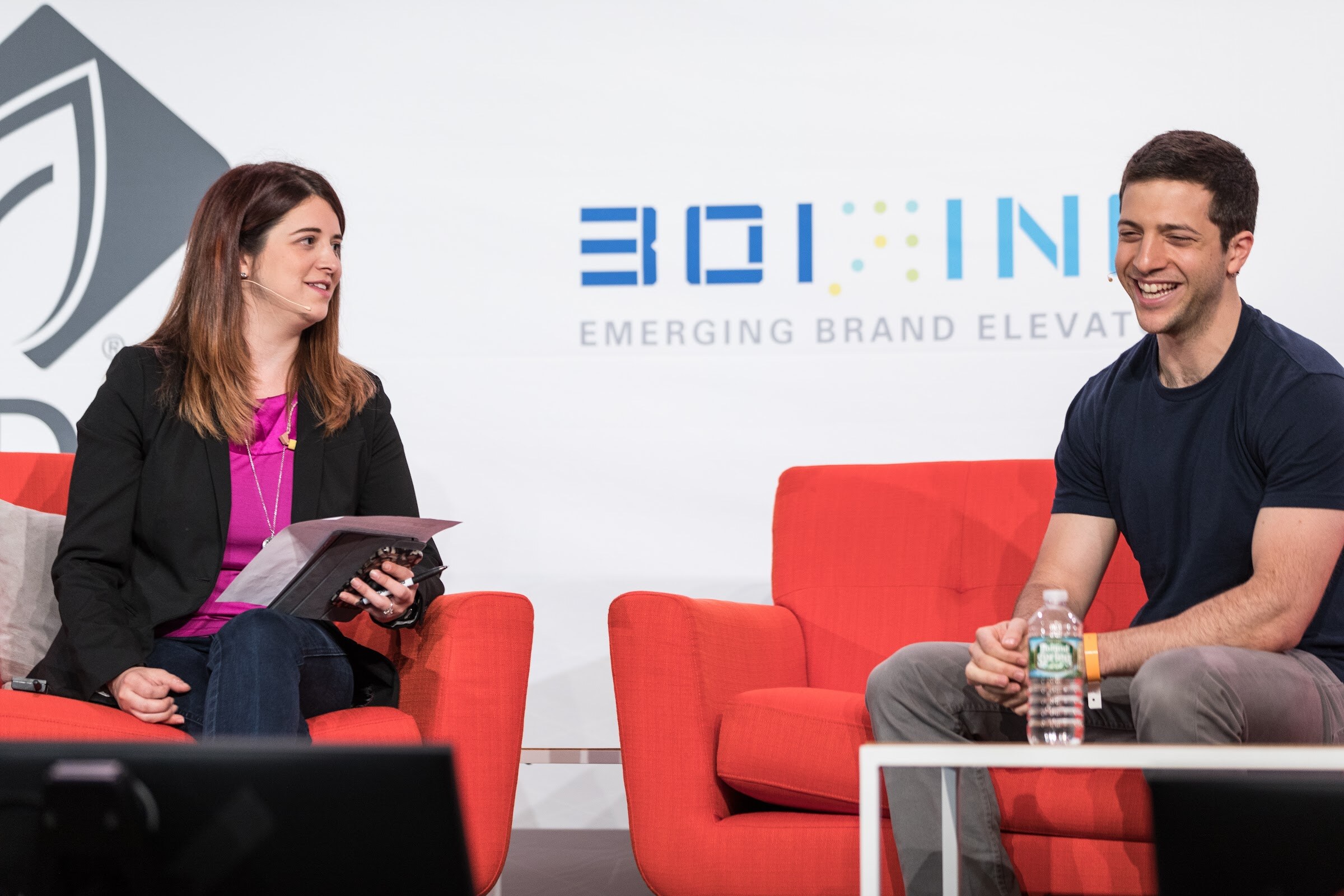 A woman in a black blazer and magenta top holds a tablet and smiles at a laughing man in a navy t-shirt in red armchairs, with a 301 Inc banner behind them.
