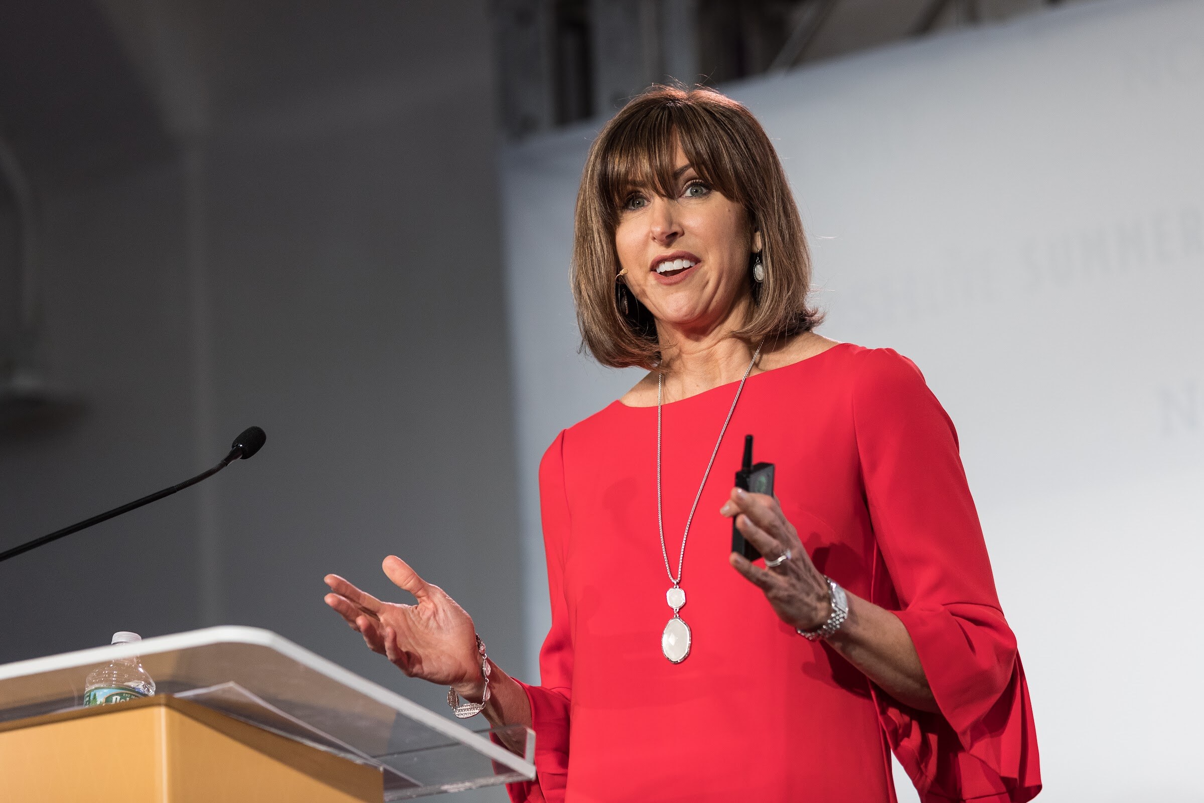 A woman with a brown bob in a red three-quarter-sleeve top speaks at a clear podium with one hand open and a clicker in the other.