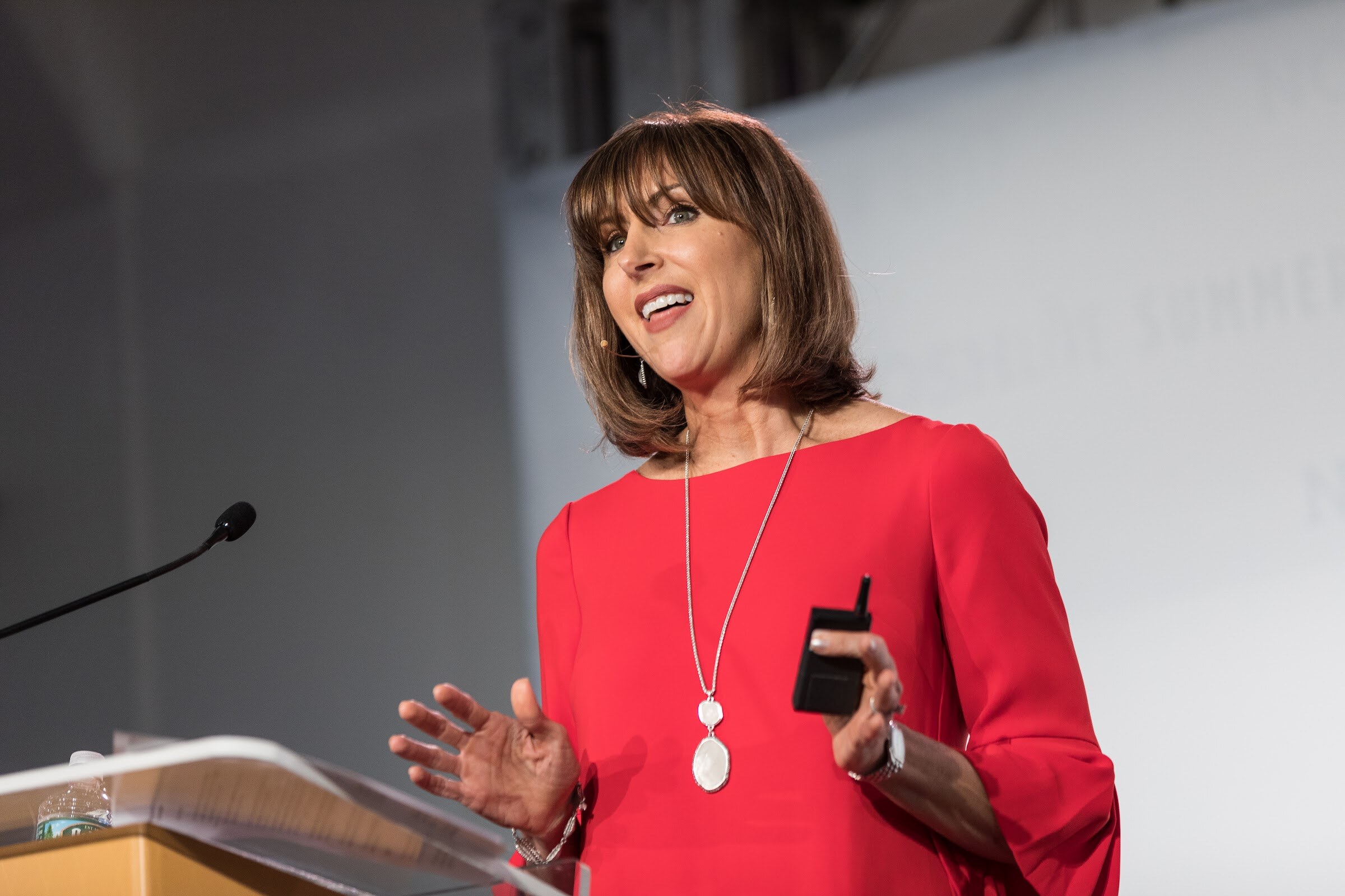A woman in a red top with a long pendant necklace smiles and speaks at a podium, holding a clicker.